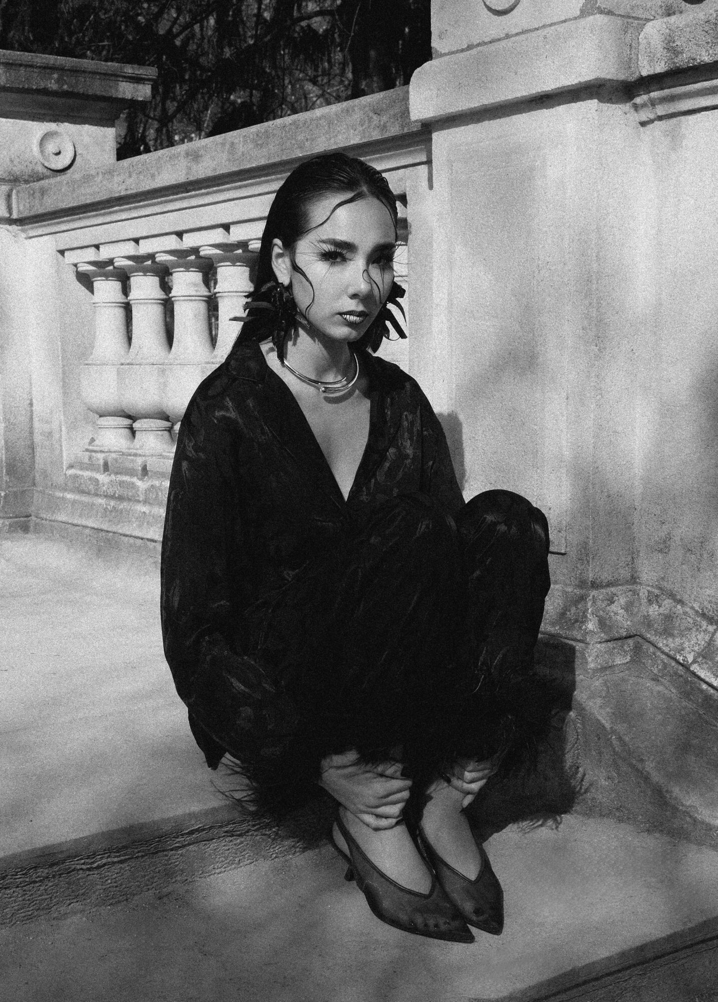 Woman crouching on ground near a stone railing, wearing dark clothing and heels, in black and white photograph.