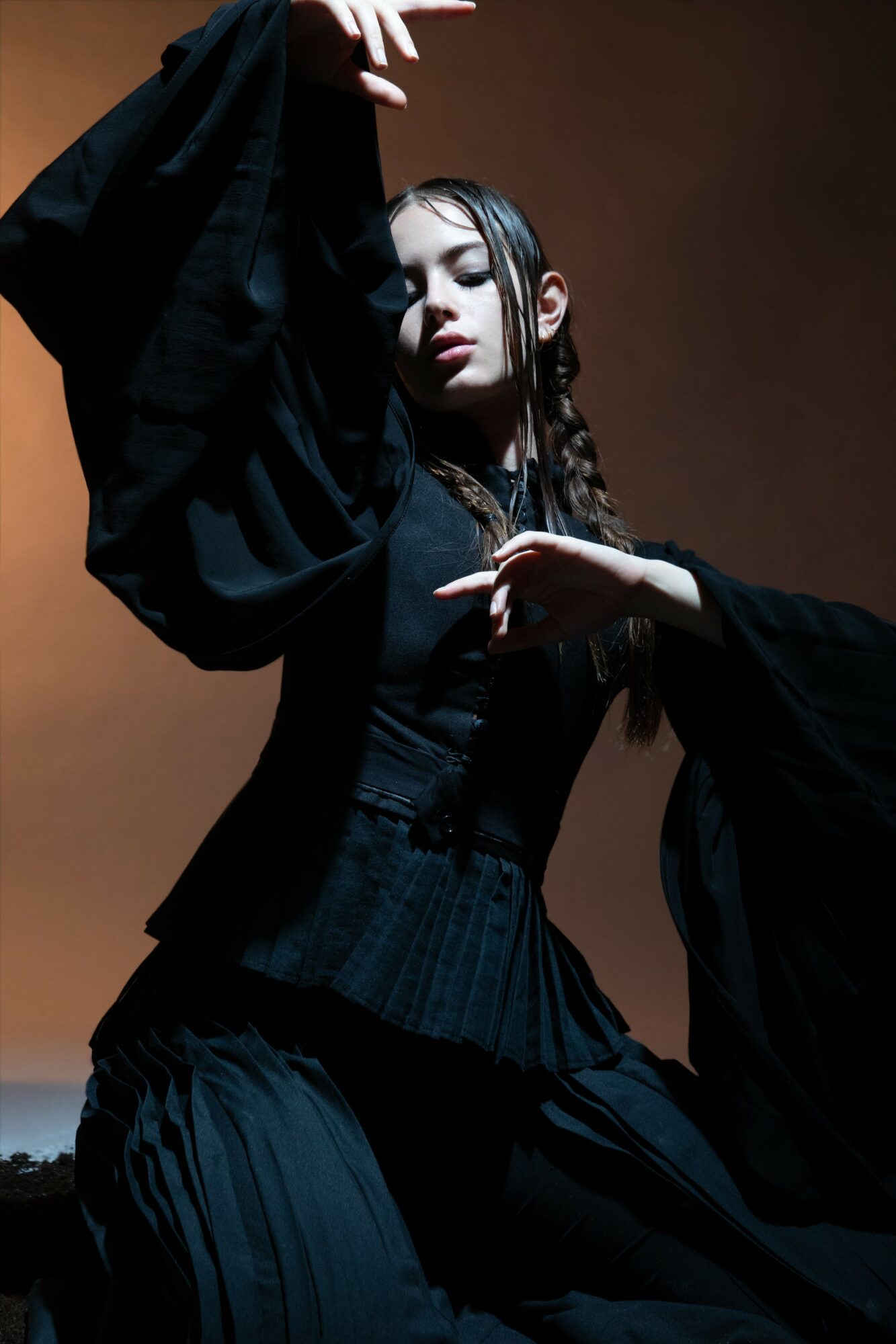 Young woman with braided hair in dark clothing poses with arms raised and eyes closed against a brown background.