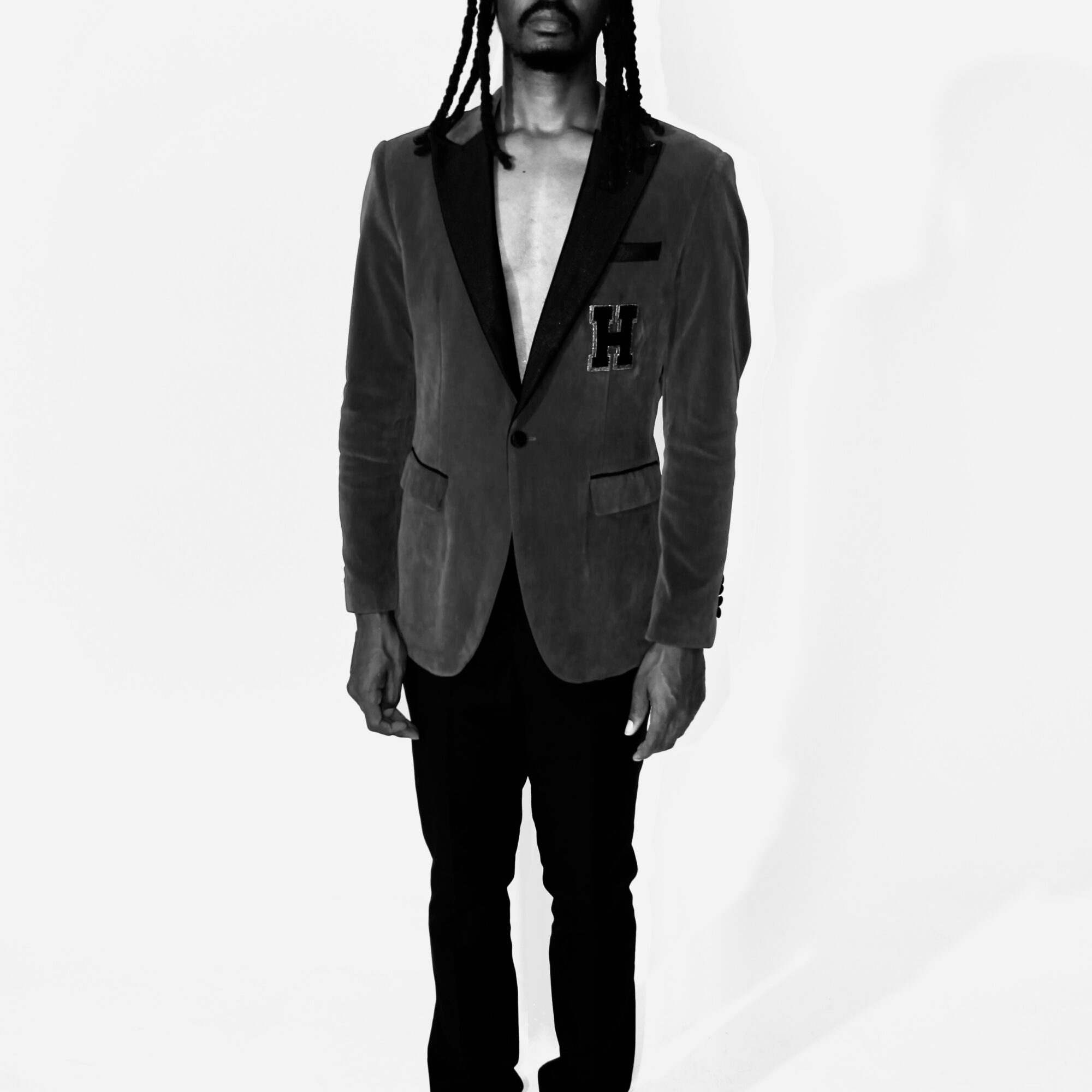 Person with long dreadlocks wearing a blazer and dark pants, standing against a plain background.