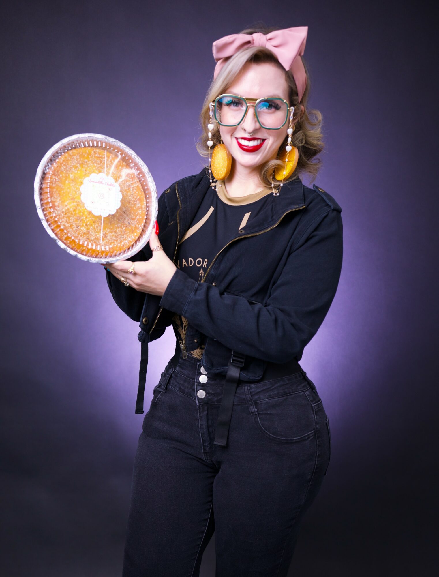 Woman with glasses, pink bow, yellow earrings, holding a round cake, smiling, against a dark background.