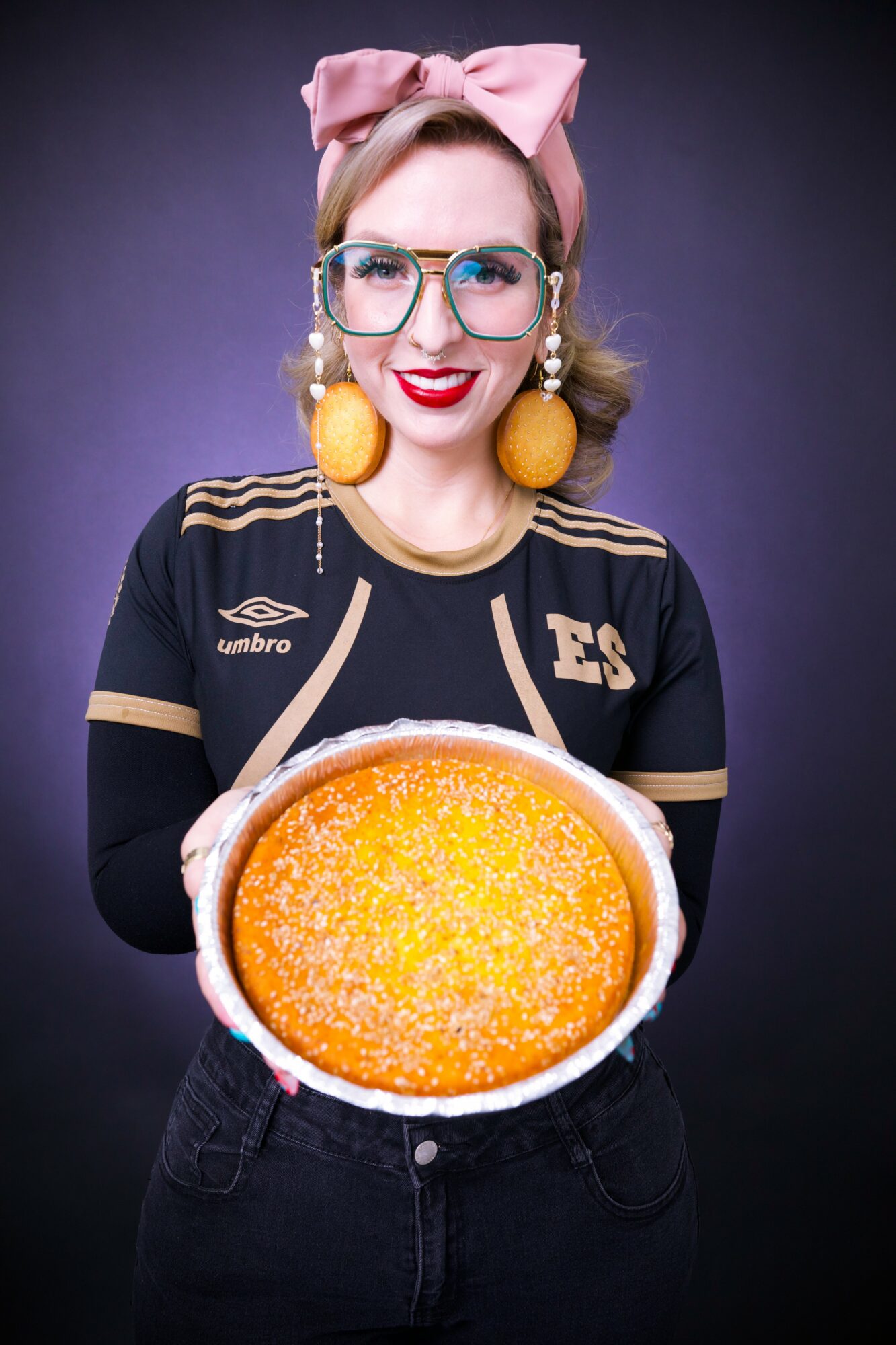 Woman with glasses, pink bow, and yellow earrings holds a large orange cake, smiling against a dark background.