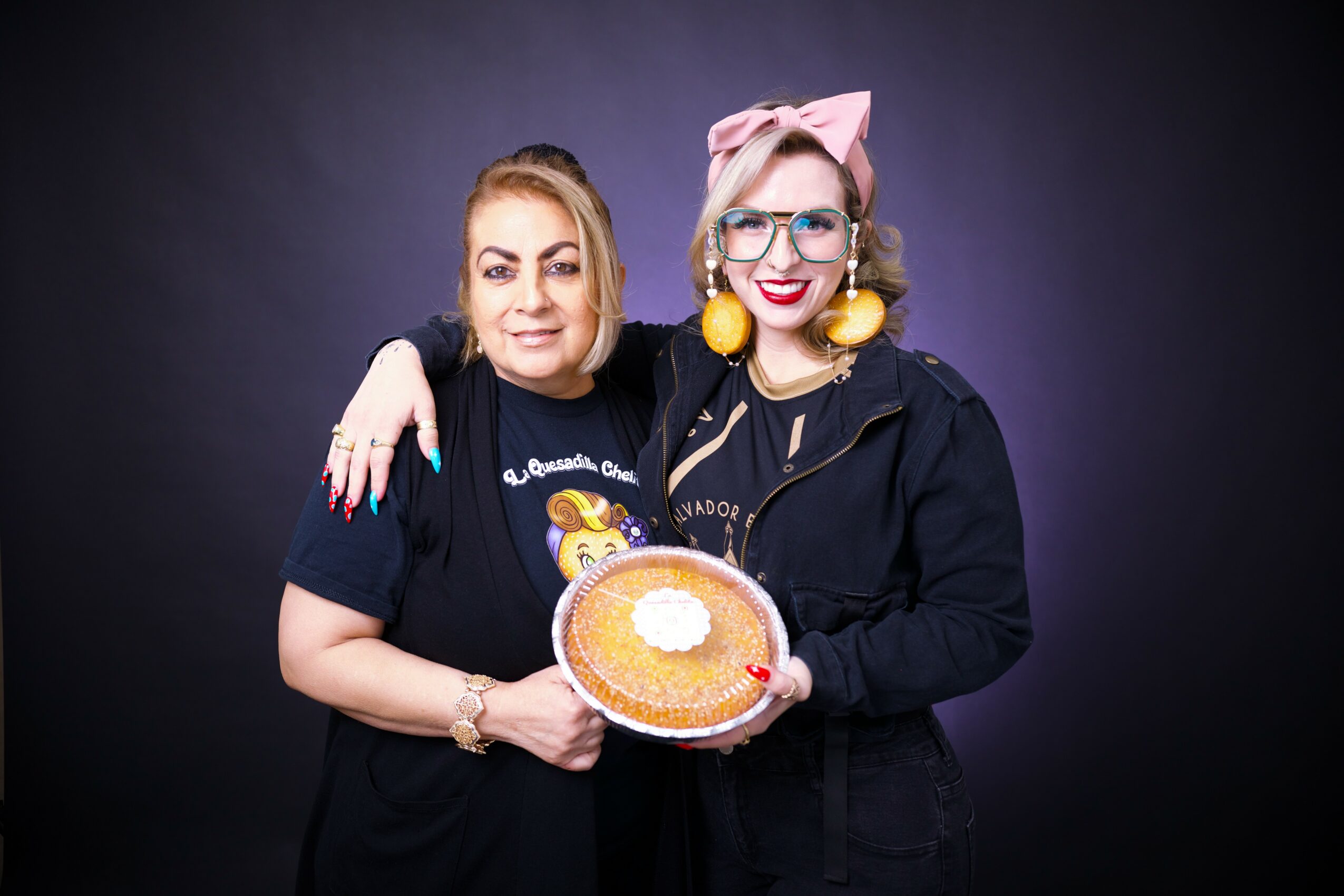 Two women smiling, holding a pie, standing against a dark background. One wears a black shirt, the other a black jacket with pink bow and yellow earrings.
