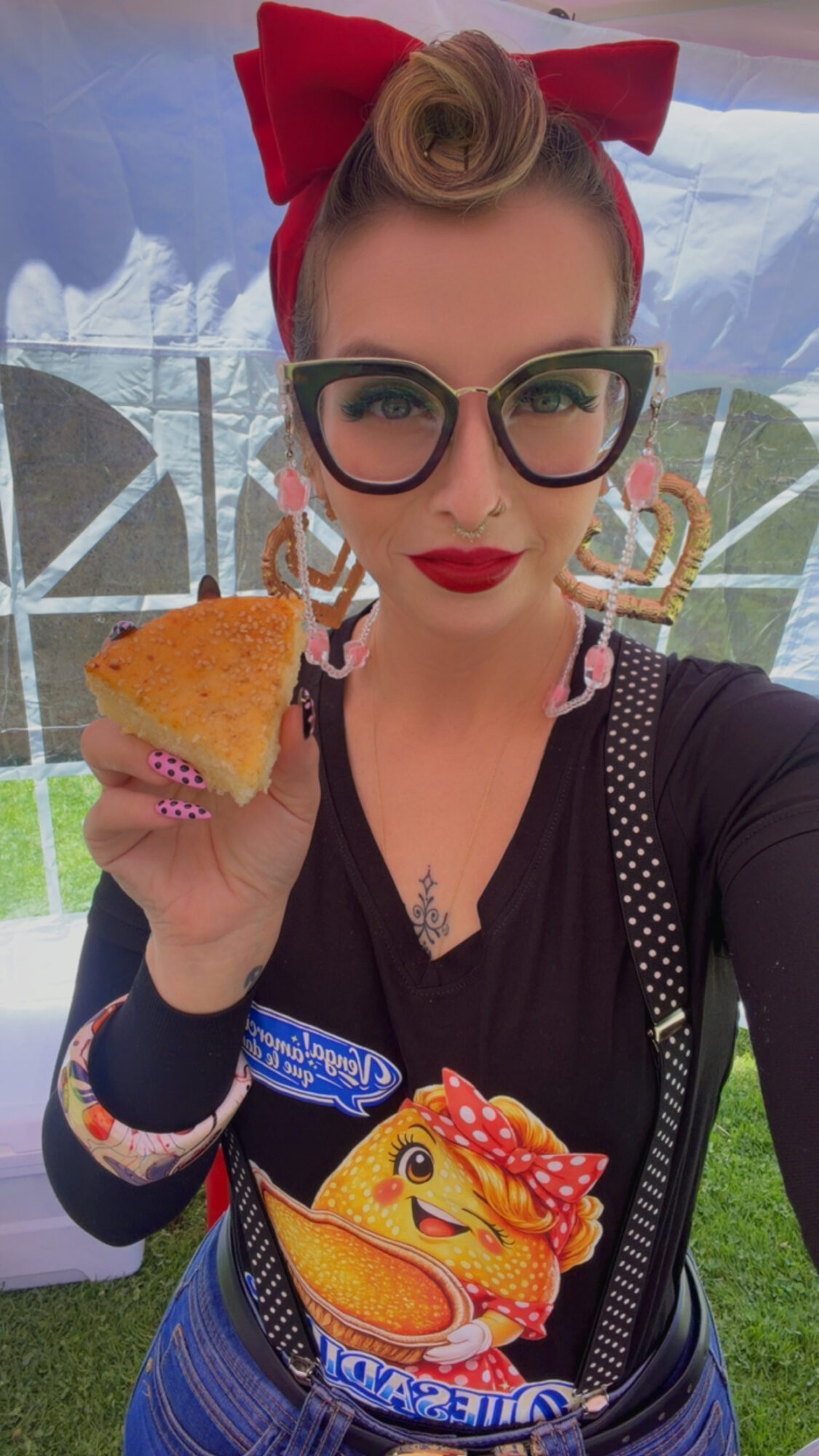 Woman with vintage hairstyle, large glasses, red bow, holding a slice of cake, wearing a colorful apron, outdoors.