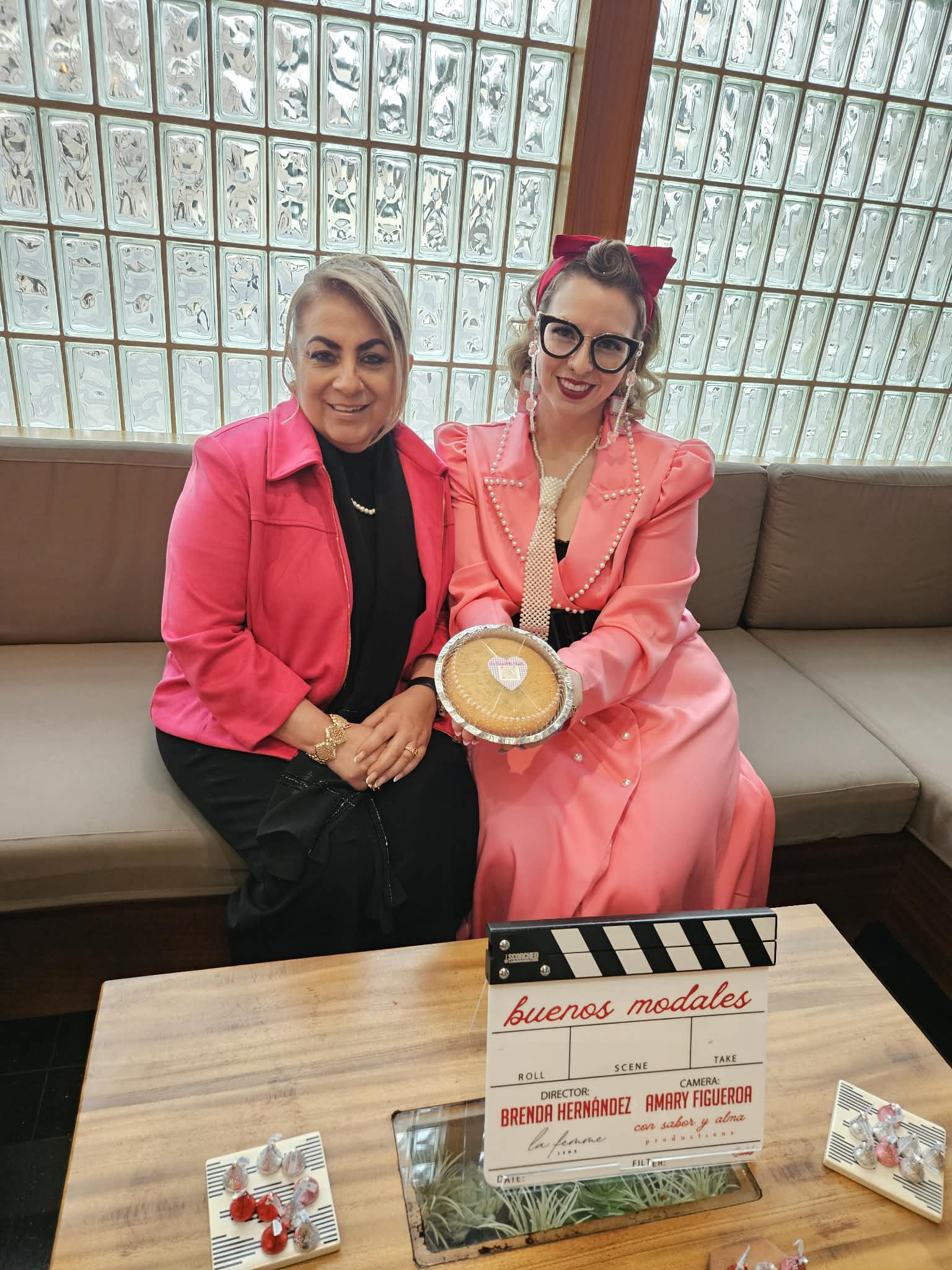 Two women sitting on a sofa, one in a pink blazer and the other in a pink dress with glasses, holding a bowl. Glass block window background.
