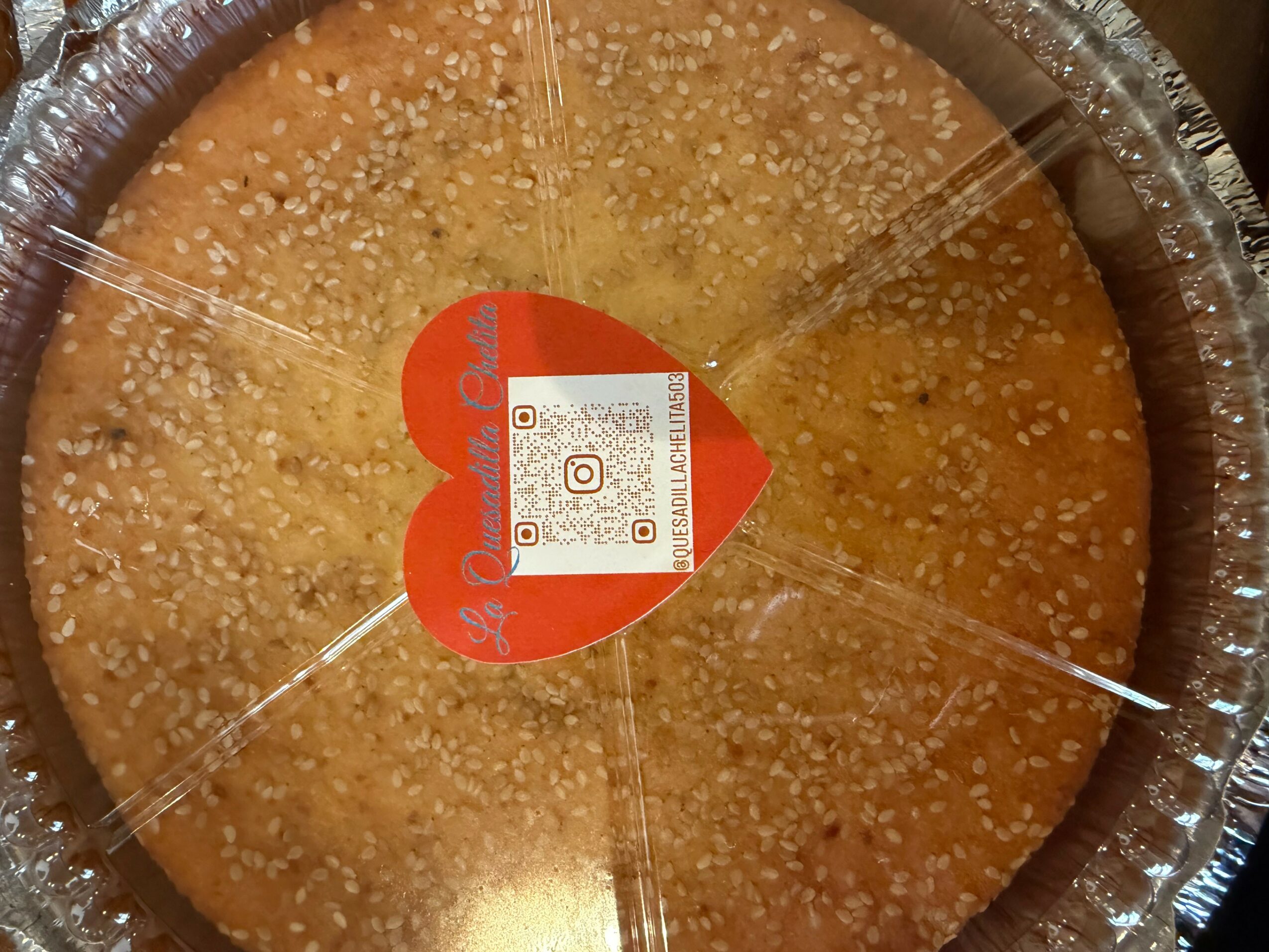 Round baked item with sesame seeds in a foil container, label with QR code and red heart shape in the center.