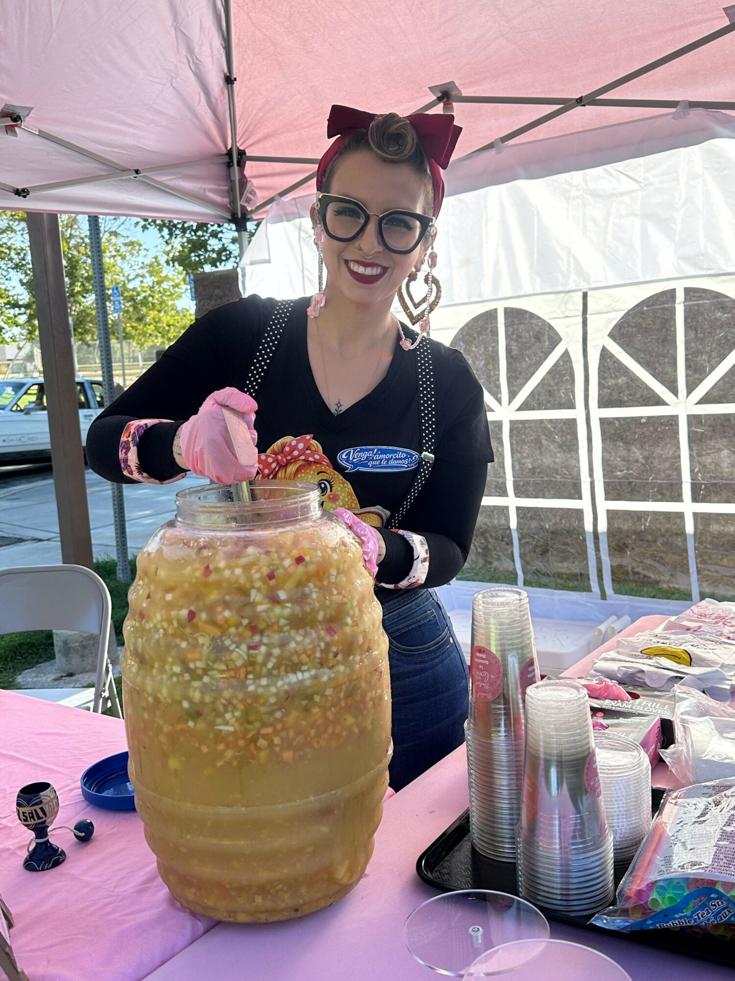 Woman with glasses and earrings, wearing a black top, stands behind a table with a large yellow cake decorated with pink flowers and sprinkles, under a pink canopy.