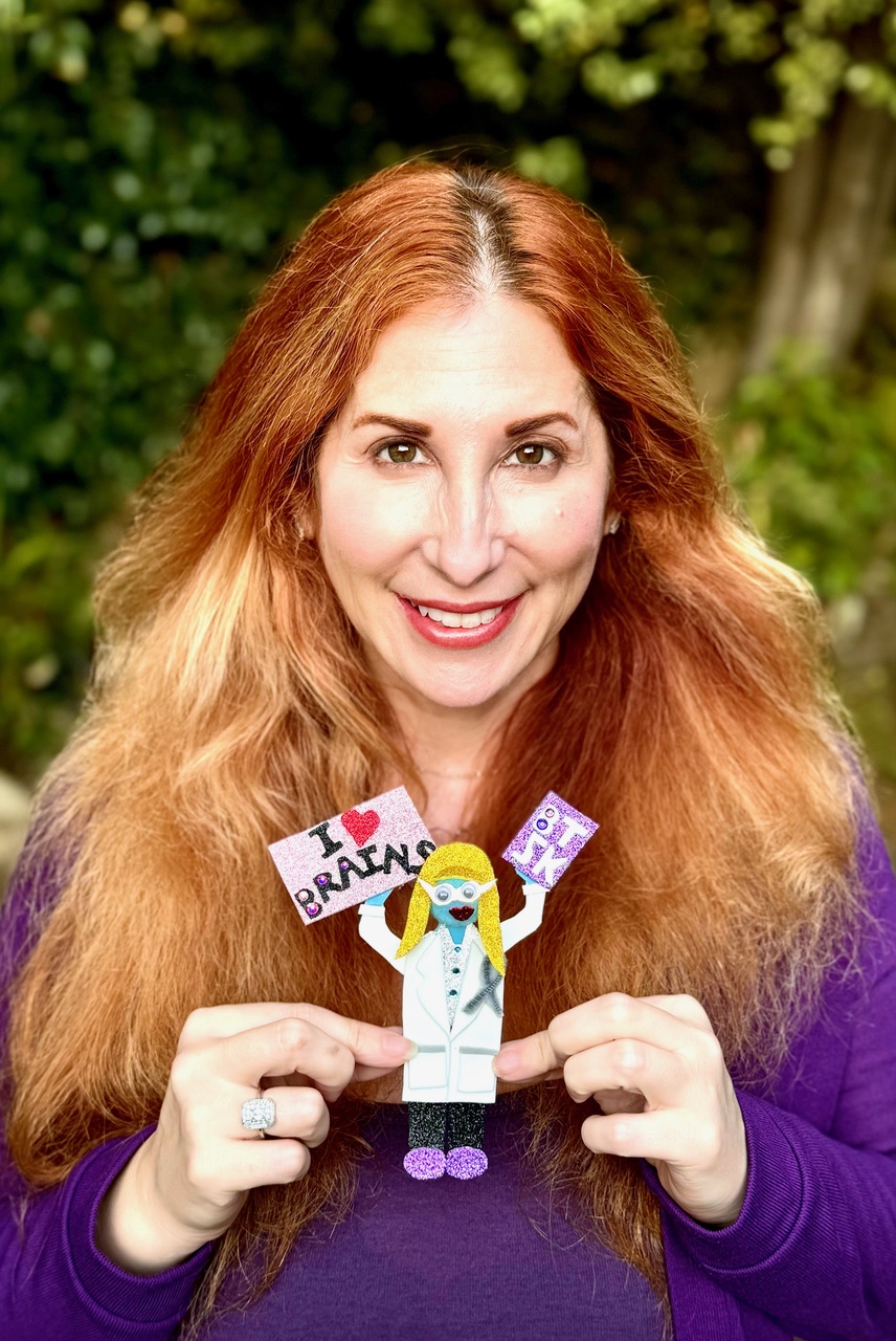 Woman with long red hair smiling outdoors, holding a paper craft of a scientist with a lab coat and glasses, background of trees.