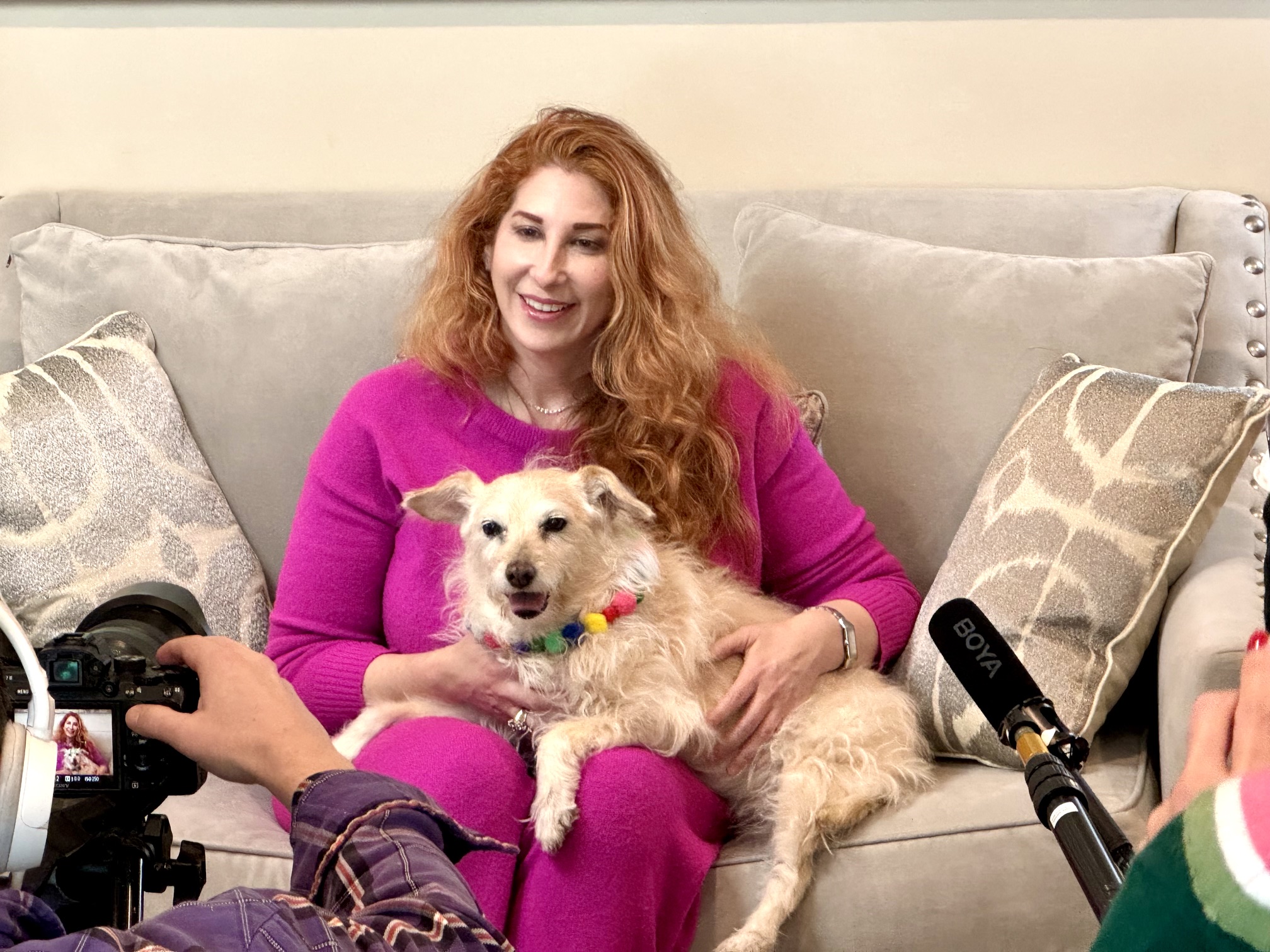 Woman with curly red hair sitting on a sofa holding a small dog, smiling, with pillows behind her.