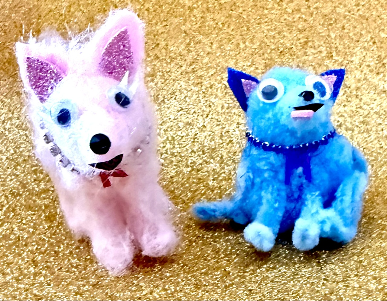 Two plush toys, one pink and one blue, sit on a gold surface, with the pink dog on the left and the blue cat on the right.