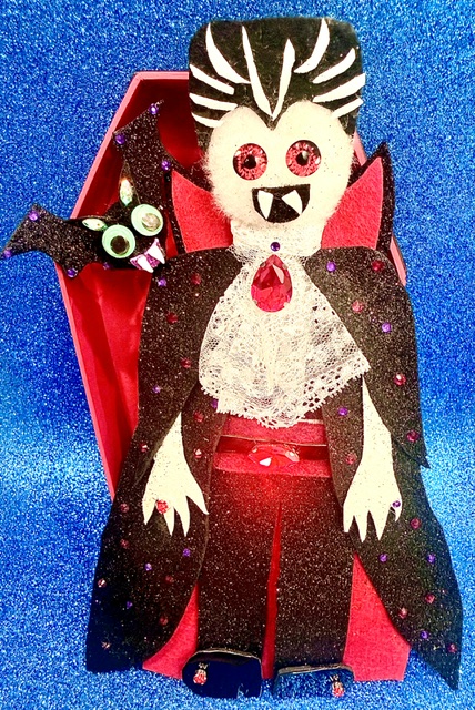 Decorative vampire figure with bat, in a red box, against a blue background, with glittery details and cartoonish features.
