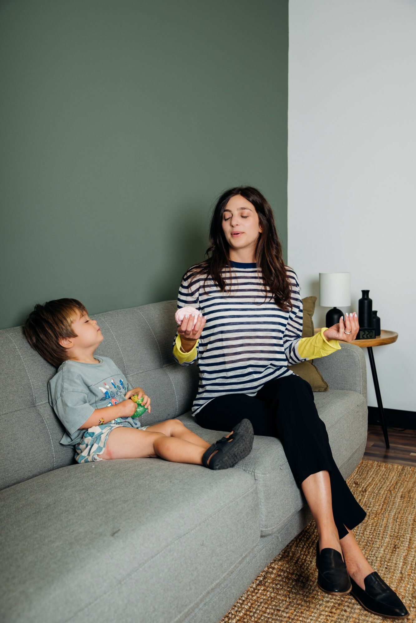 Woman with eyes closed, sitting on sofa, child sitting beside her, both in casual clothing, room with green and white walls.