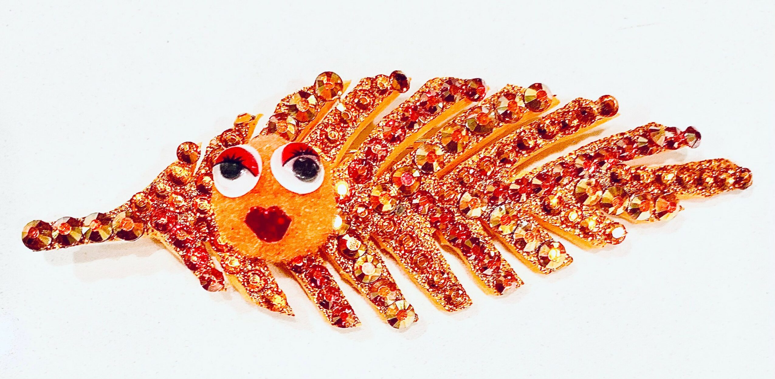 Lobster-shaped decoration made of orange and red rhinestones with googly eyes and a small mouth, on a white background.