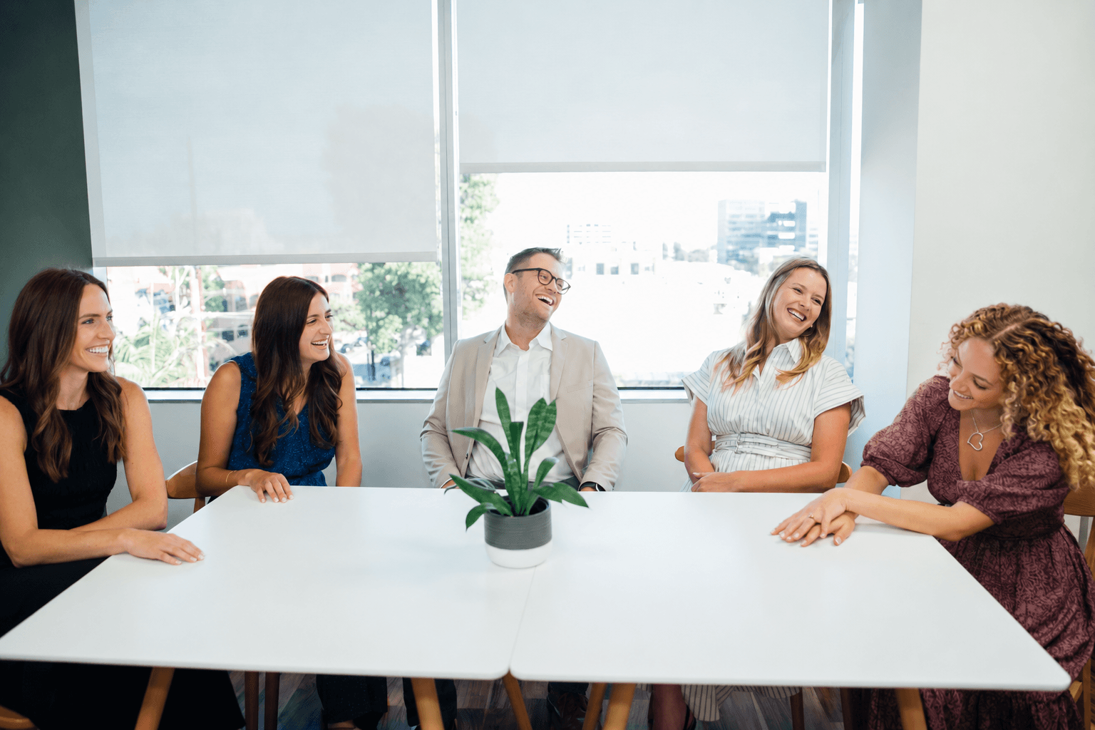 Five people sitting at a white table in a bright room with large windows, smiling and talking, with a plant in the center.