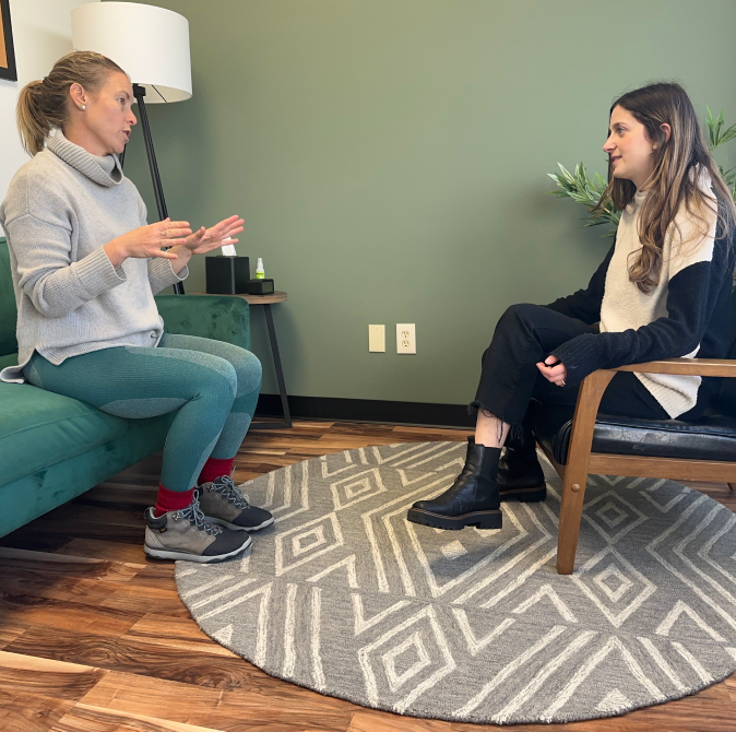 Two women sit facing each other in a room with a green wall, a patterned rug, and wooden flooring.