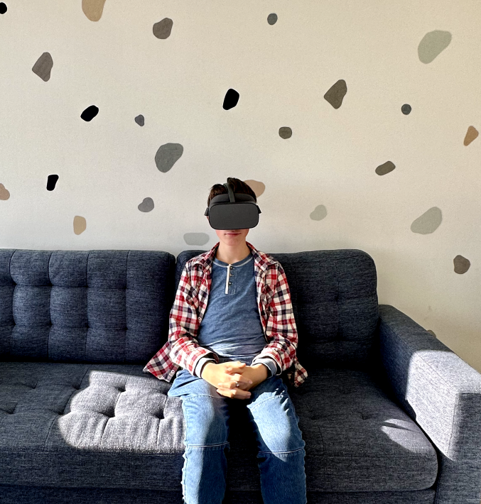 Person sitting on a dark gray sofa wearing a virtual reality headset, plaid shirt, and jeans, with a patterned wall behind.