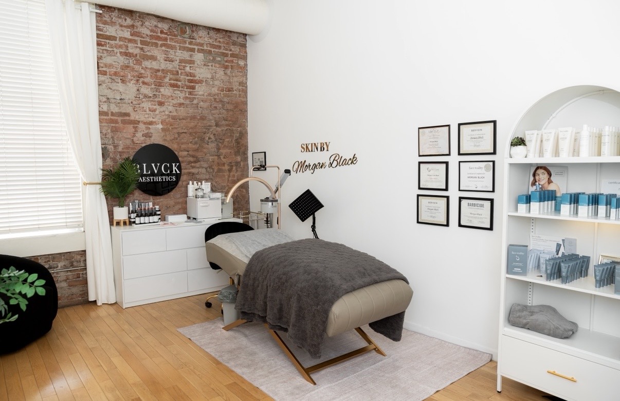 Massage table with gray blanket in a room with white walls, brick accent wall, and shelves with towels and products.