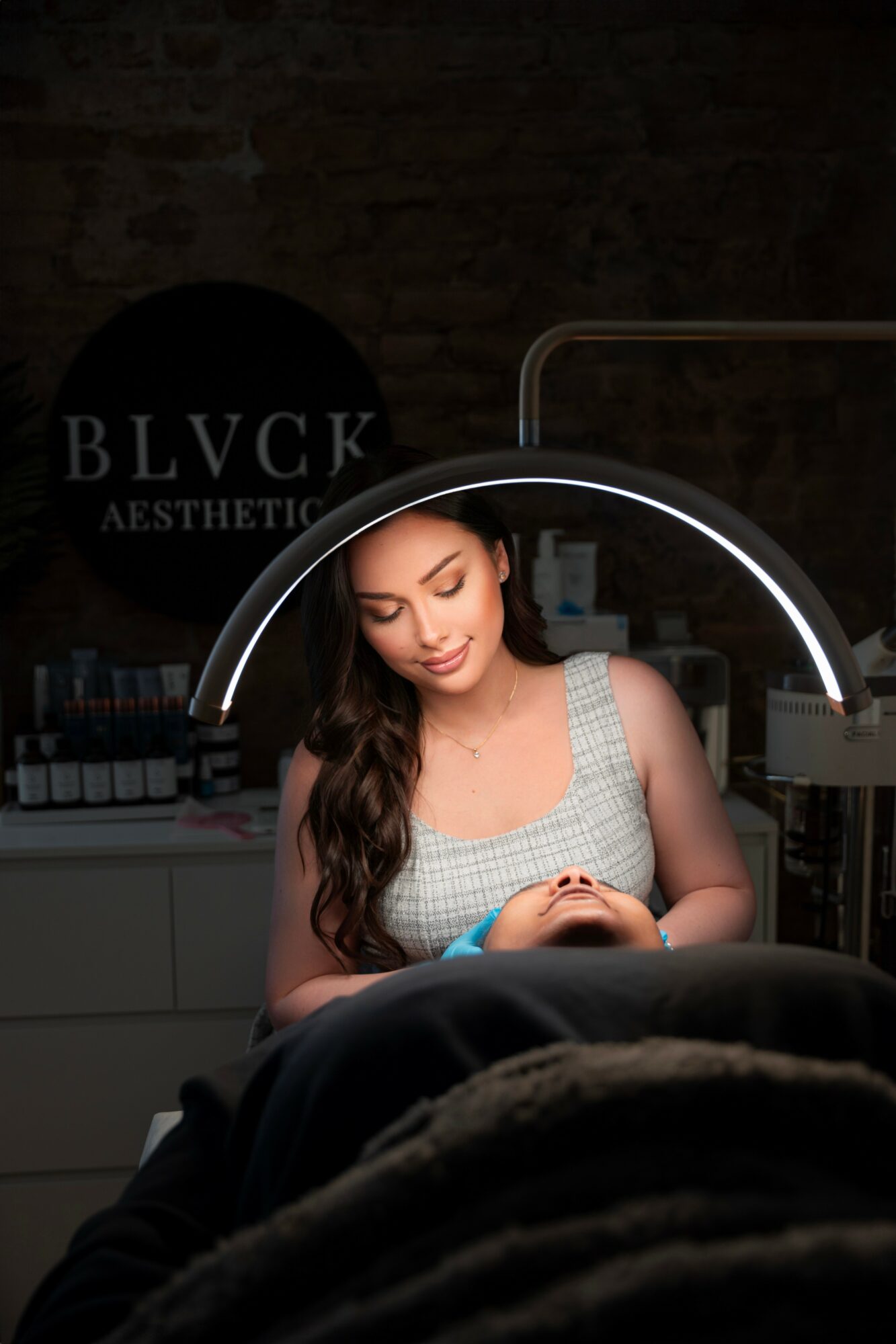 Woman with long dark hair receiving a manicure under a ring light in a salon, with a sign reading BLVCK in the background.