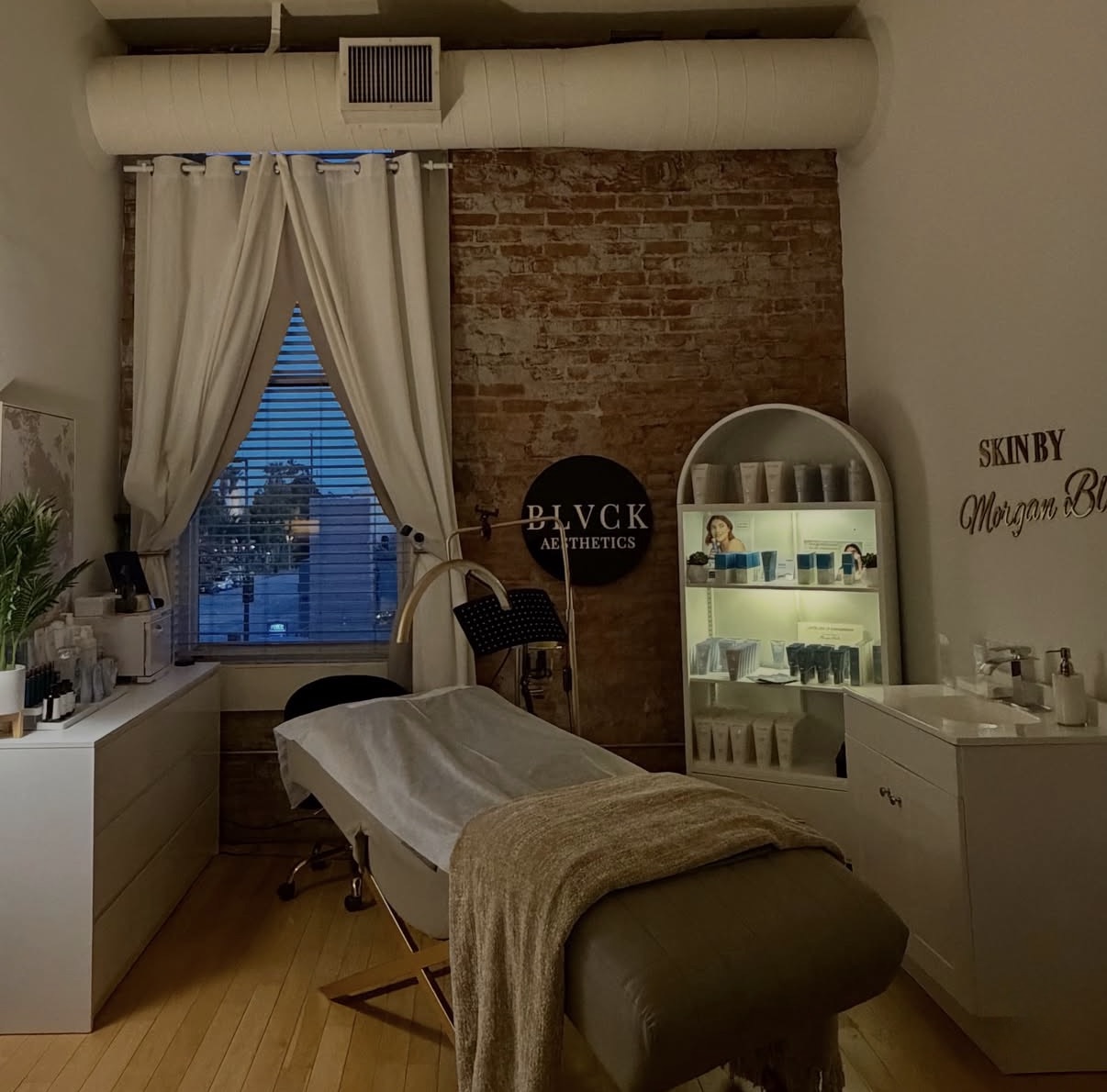 Treatment room with massage table, window with curtains, brick wall, and shelves with products.