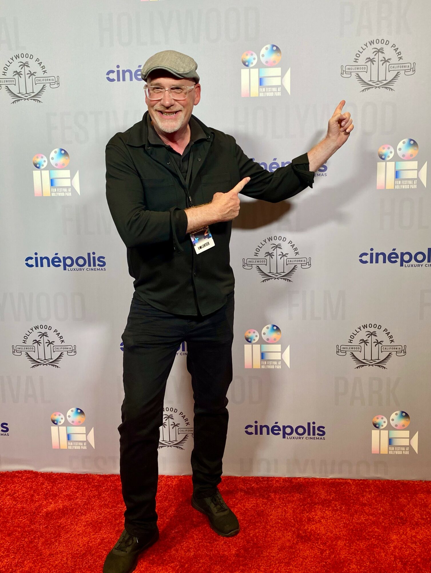 Man in black clothing and a cap smiling and pointing at a backdrop with logos, standing on a red carpet.