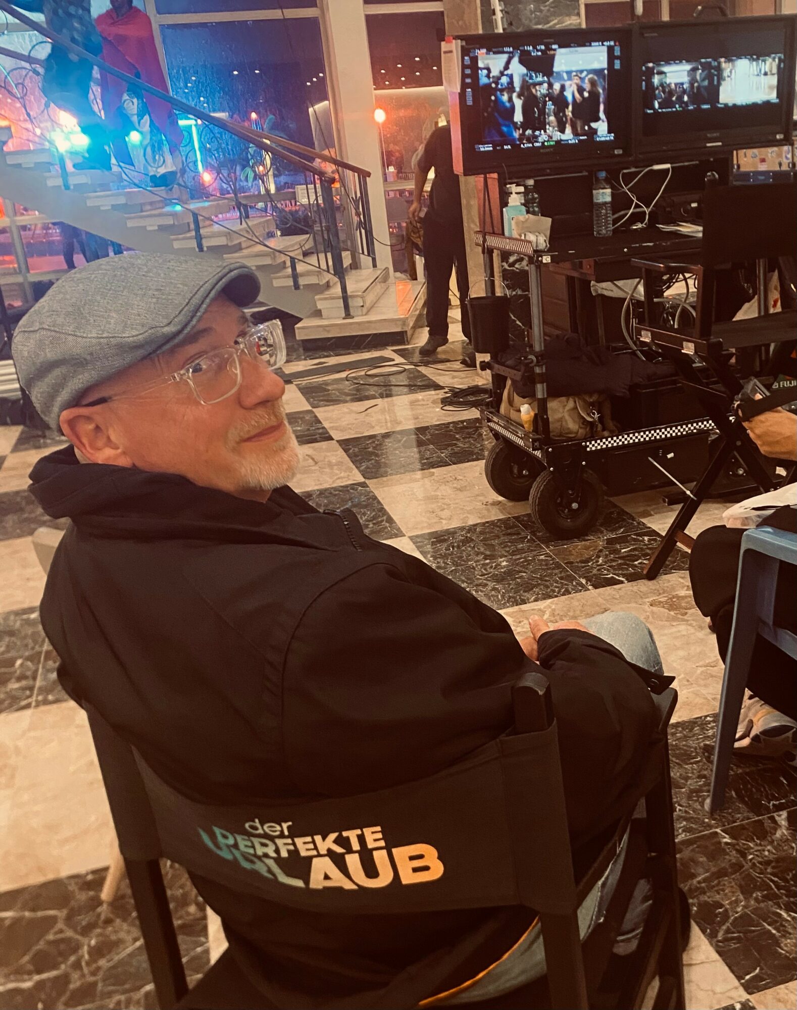 Man wearing glasses and a gray cap sitting in a chair at a film or TV set, with monitors and equipment nearby.