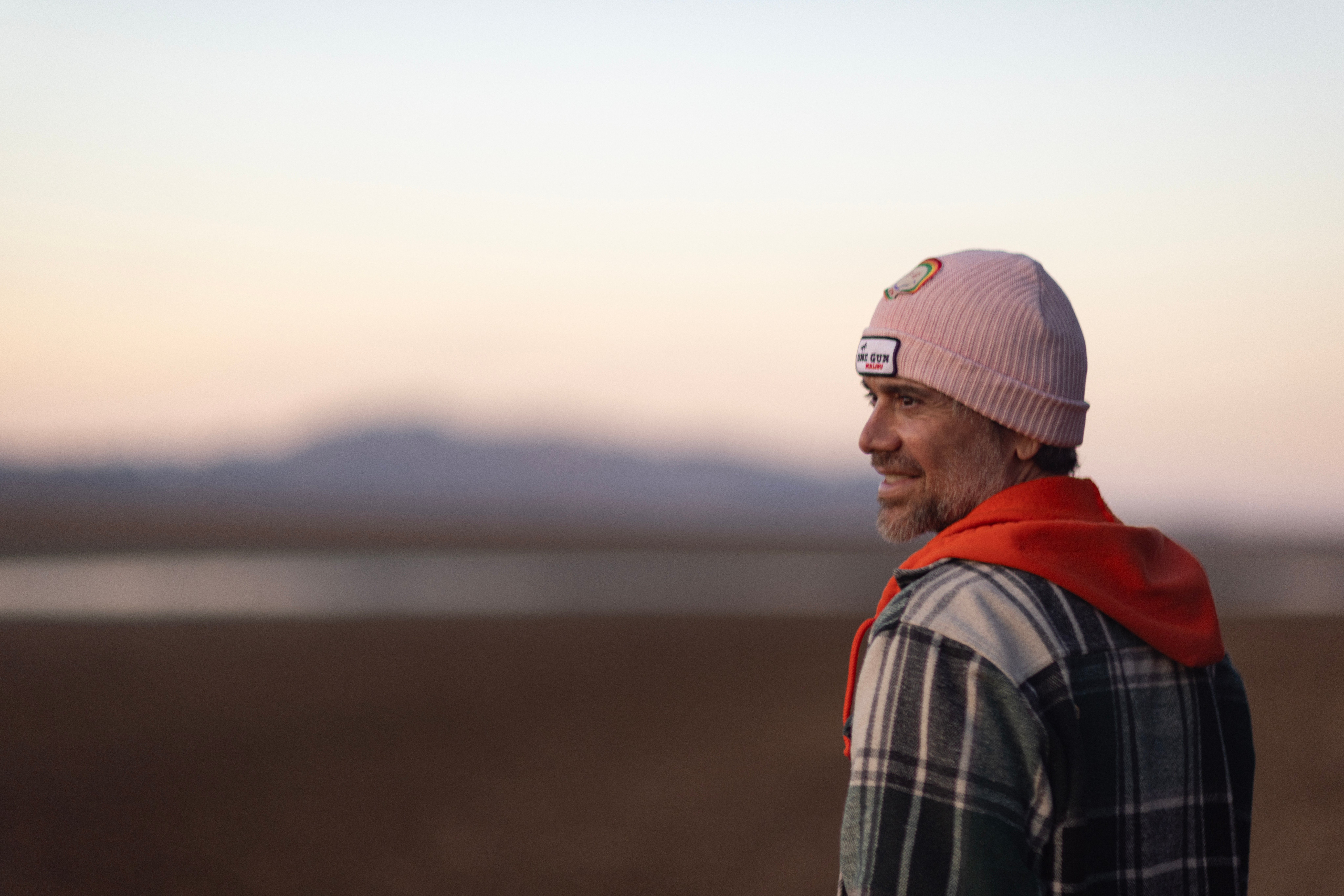 Man wearing a beanie and plaid jacket outdoors during sunset, smiling and looking to the side.