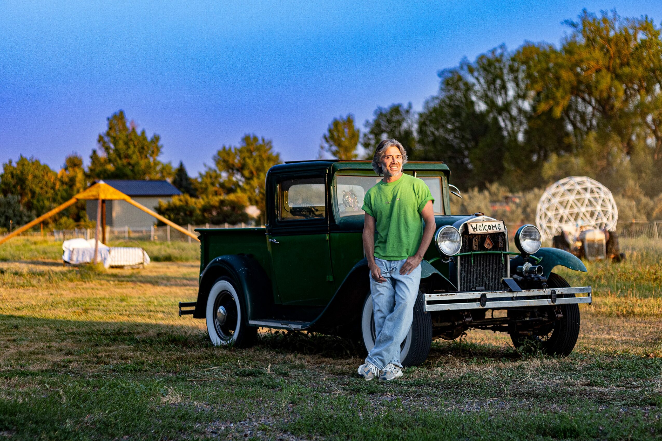 Man in green shirt leaning against a vintage black truck outdoors with trees and structures in background.