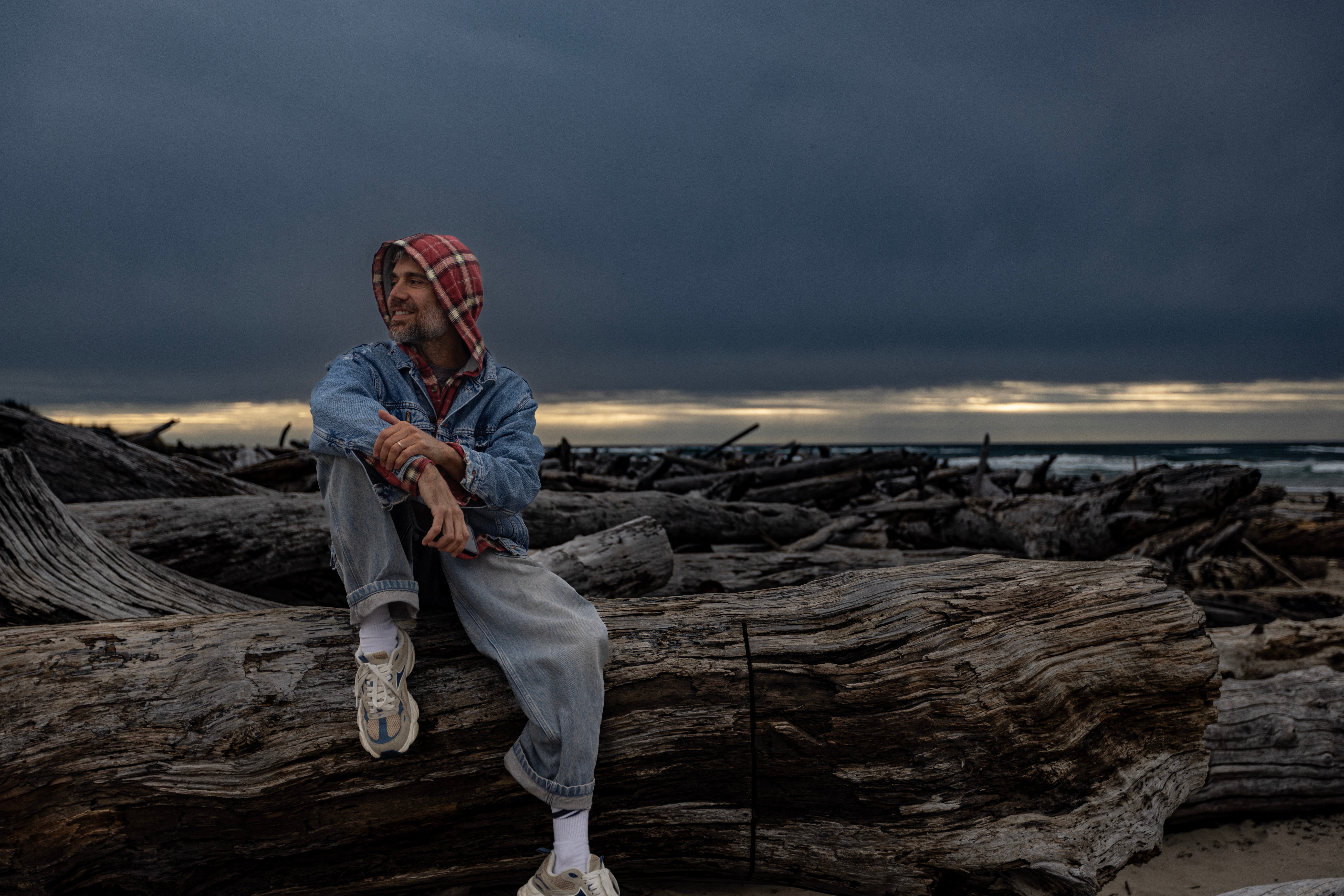 Person sitting on driftwood at the beach with dark stormy sky in background.