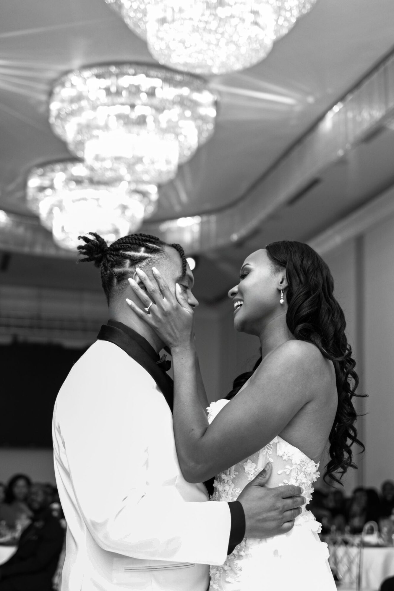 A couple dancing closely at a formal event, with chandeliers overhead and guests in the background.