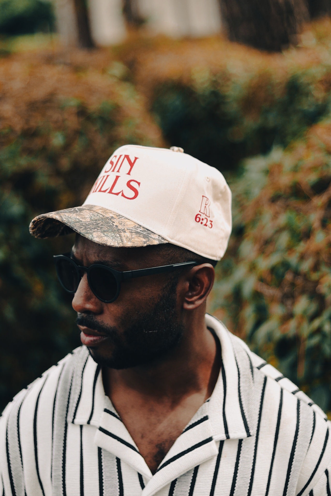 Man wearing sunglasses and a cap with text, outdoors with blurred foliage background.