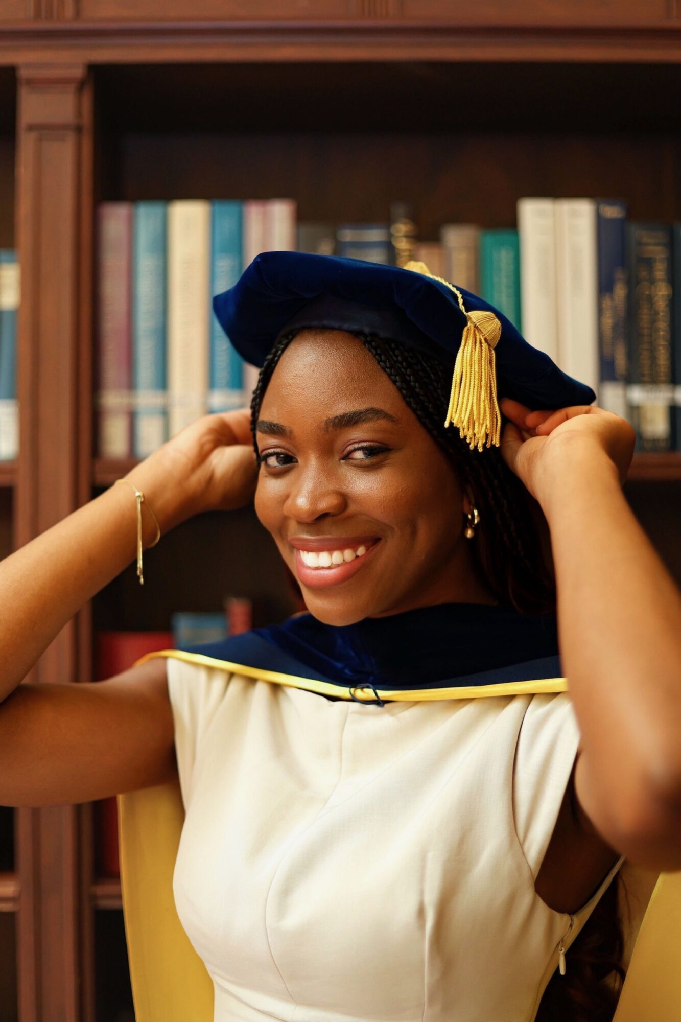 Young woman in graduation cap and gown smiling, adjusting her cap in front of a bookshelf.