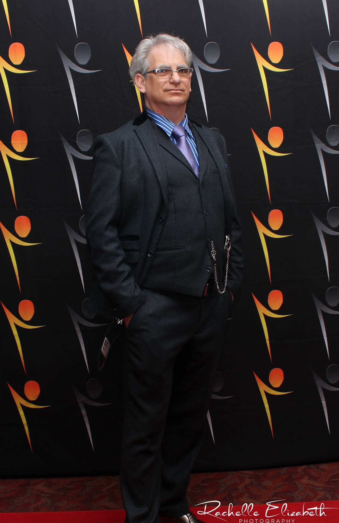 Man in a suit standing with hands in pockets in front of a patterned backdrop.