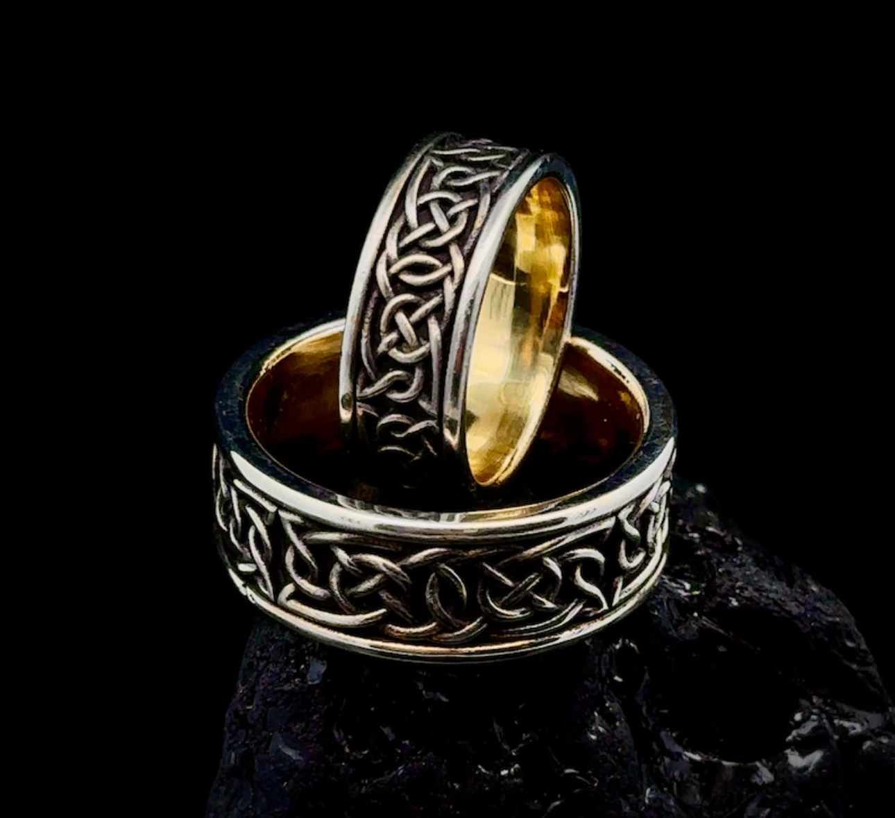 Two rings with intricate Celtic knot designs, one gold and one silver, placed on a dark surface against a black background.