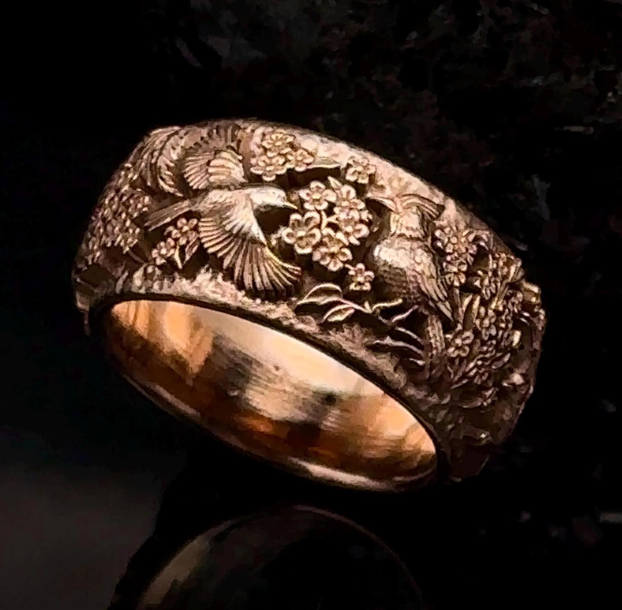 Intricately carved ring with birds and floral patterns, gold-colored, on a dark background.
