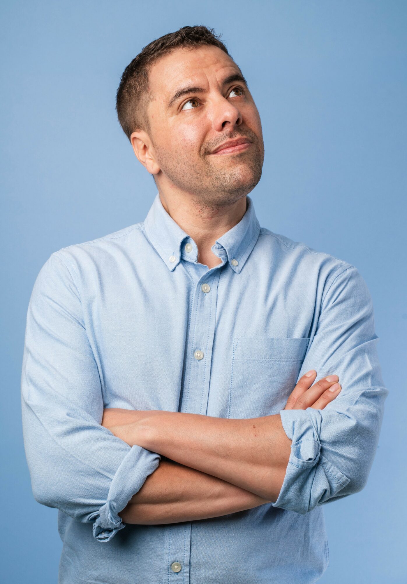 Man with short hair and a light blue button-up shirt, arms crossed, looking upward, against a blue background.