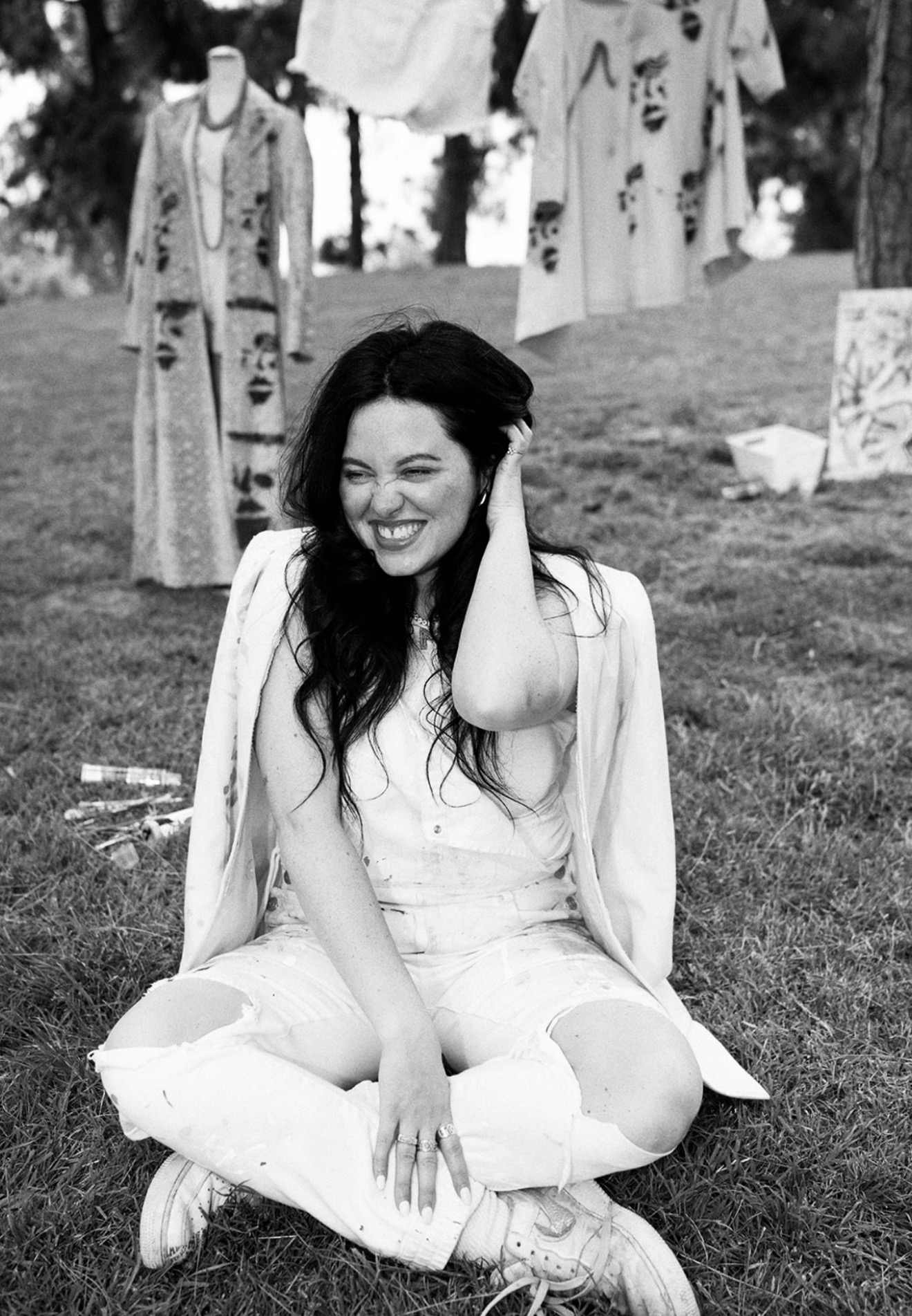 Woman sitting on grass, smiling, with hanging clothes in the background, outdoors during daytime.