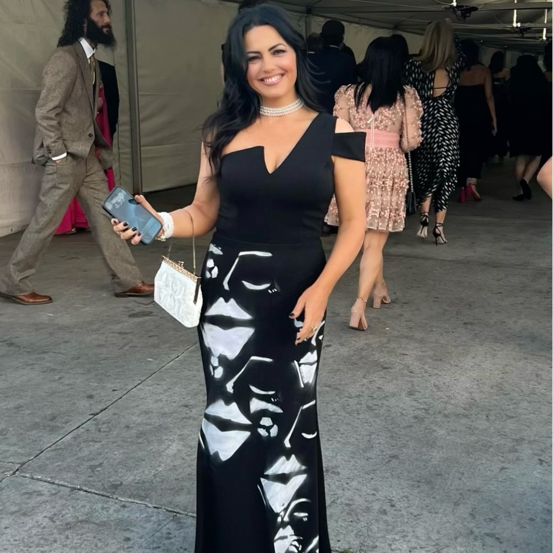Woman in black dress holding a phone and purse, smiling at an event with people in the background.