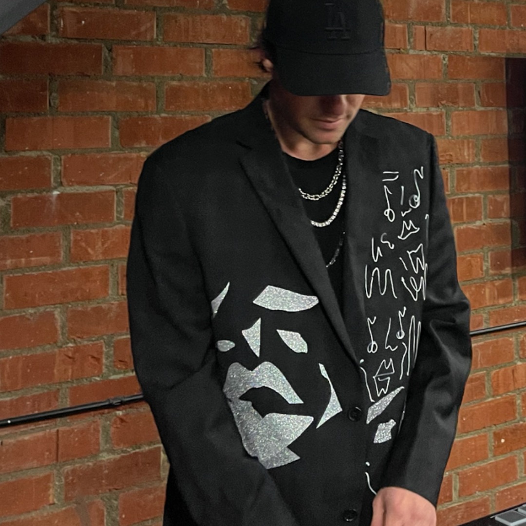 Person wearing a black jacket with white abstract designs, black cap, and layered necklaces, standing against a brick wall.