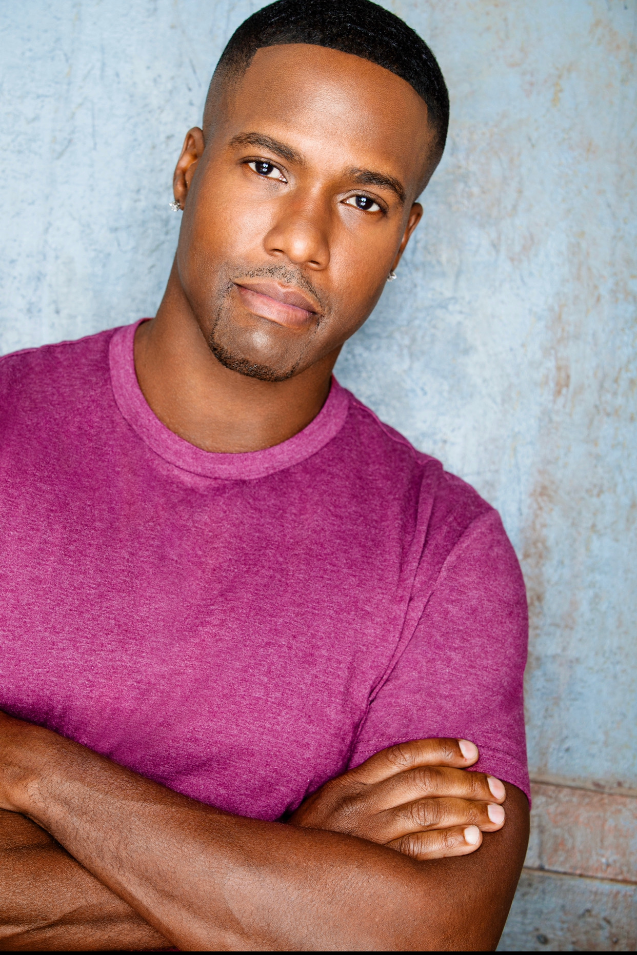 Man with short hair and a serious expression, wearing a purple shirt, standing against a textured blue background.