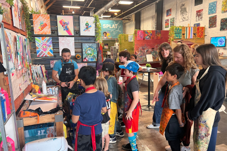 Group of children and adults gathered around an artist demonstrating art techniques at an indoor art event.