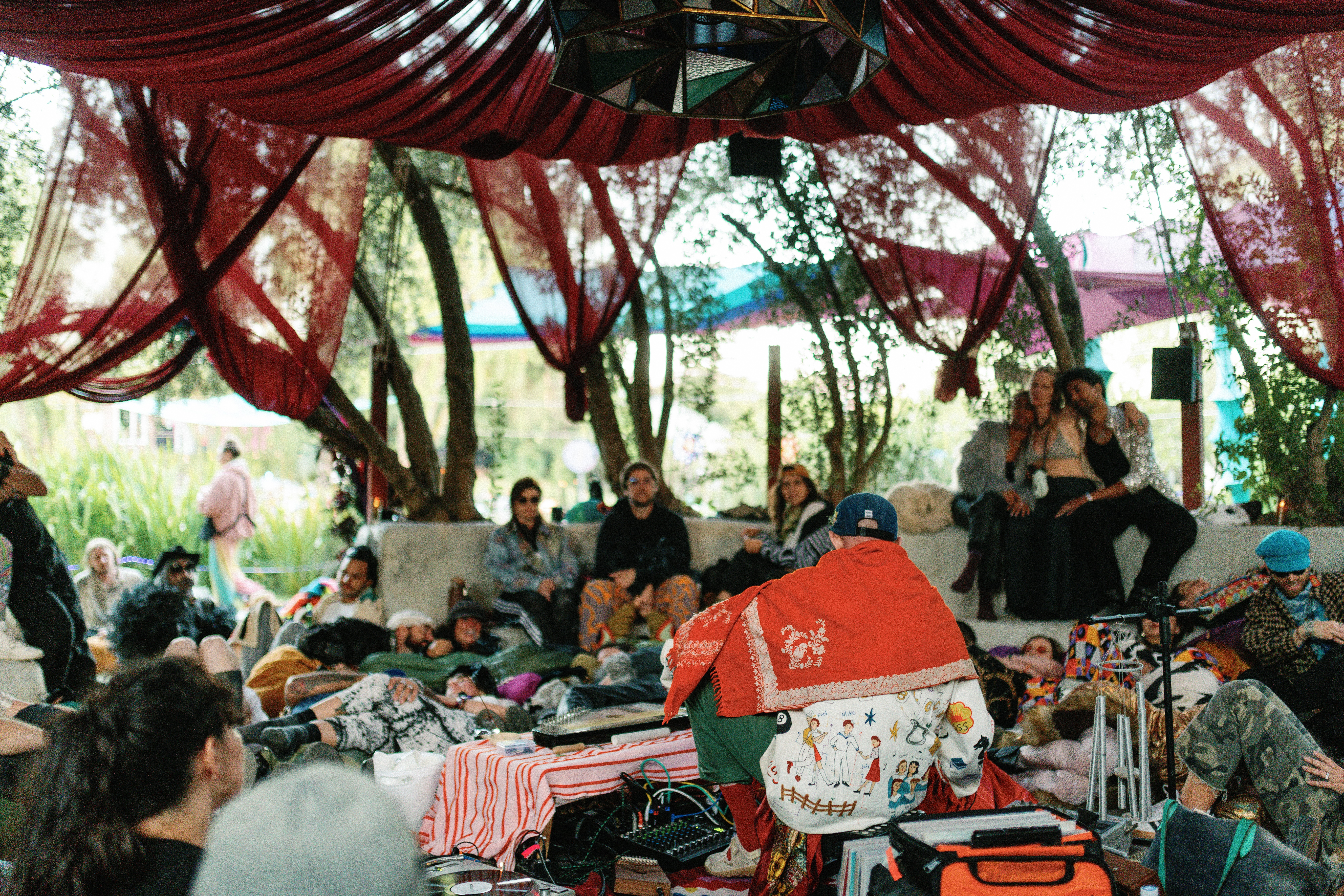 People sitting and browsing items at an outdoor market under a red canopy, with trees and sunlight in the background.