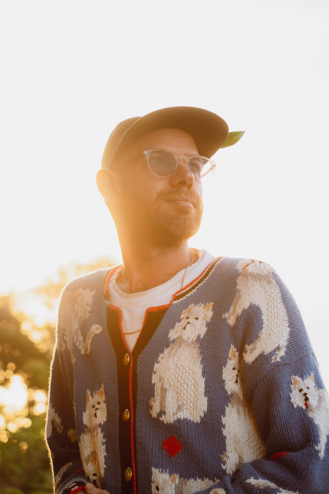 Man wearing sunglasses and a cap, outdoors, sunlight, wearing a sweater with dog patterns, looking to the side.