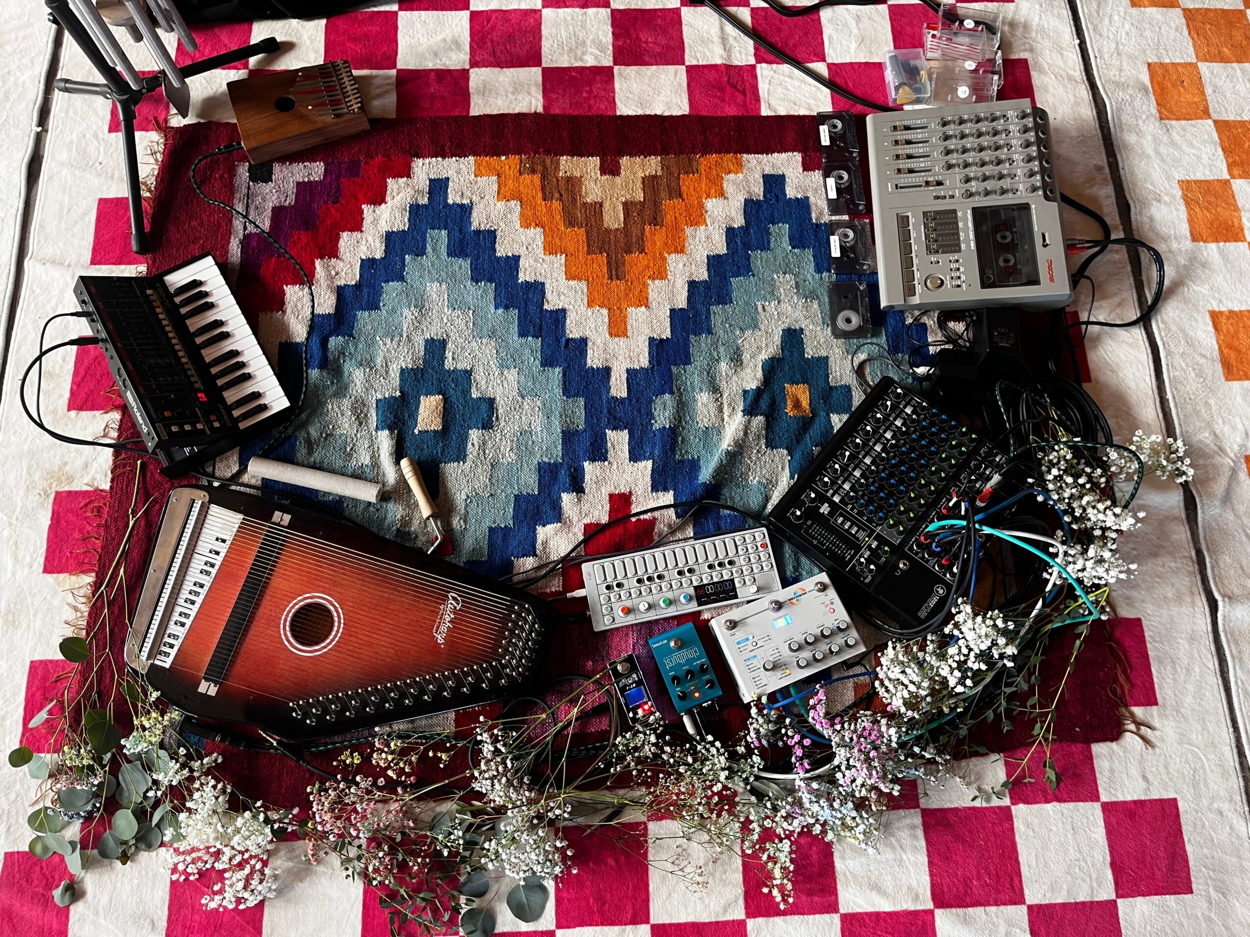 Music equipment including a keyboard, mixer, and synthesizer on a colorful patterned rug with flowers and checkered floor.