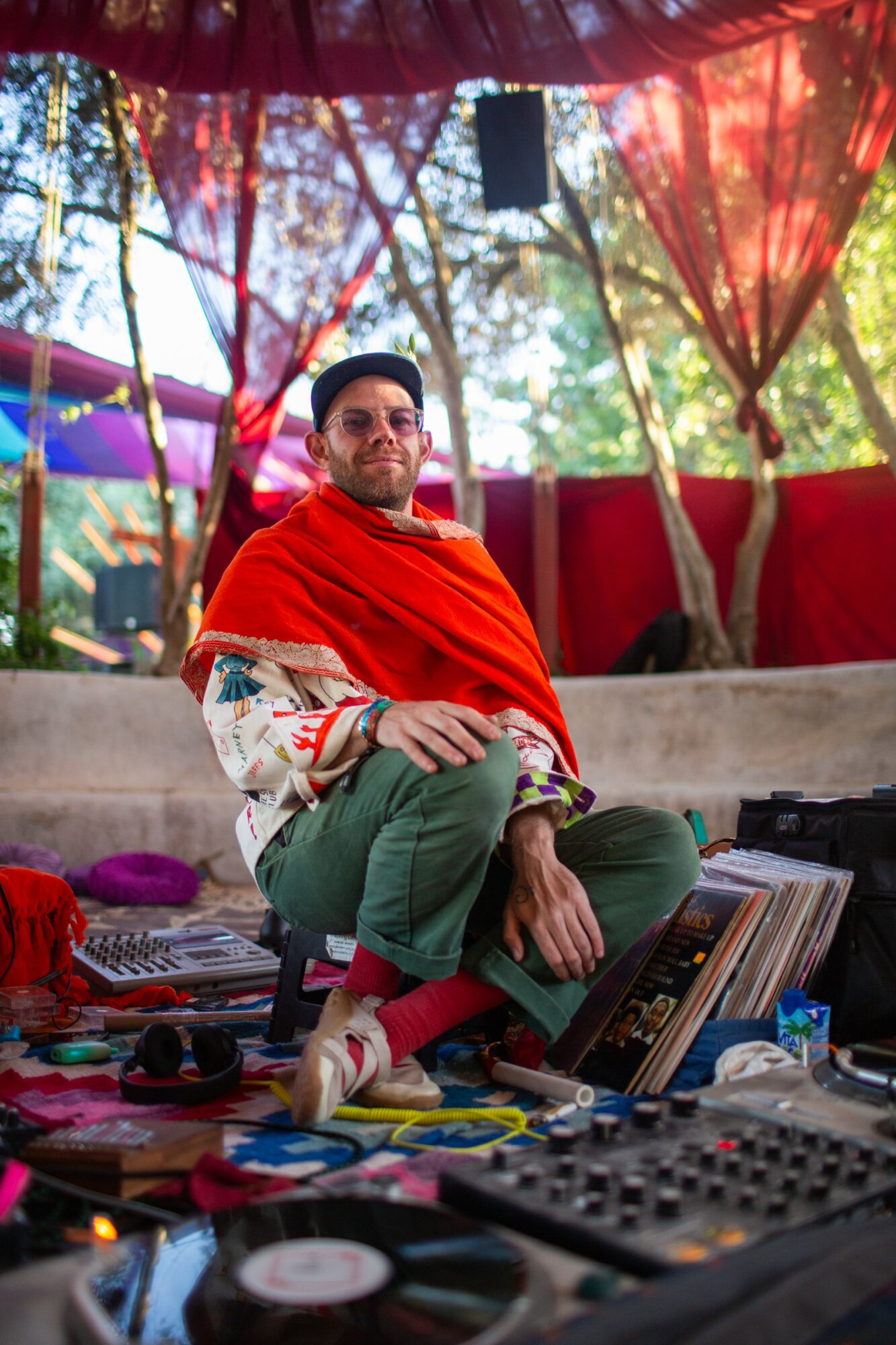 Person sitting cross-legged outdoors with colorful fabric and trees in background, surrounded by music equipment and records.