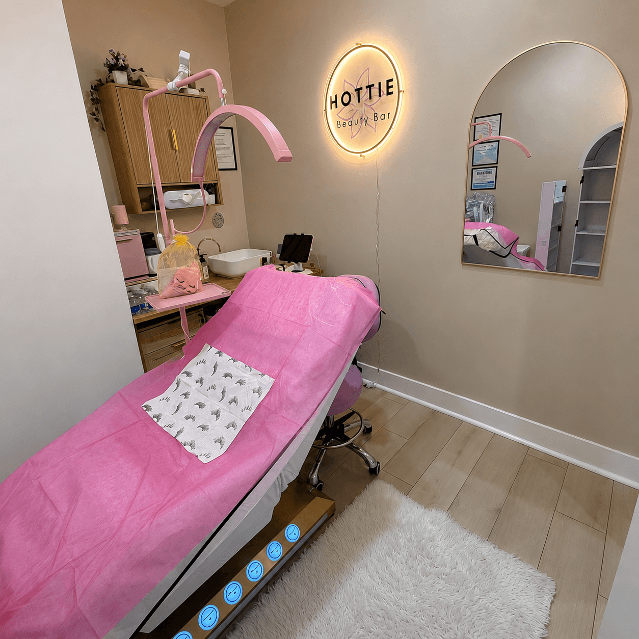 Treatment room with a pink-covered bed, mirror, and a neon sign reading 'HOTTIE' on beige wall.
