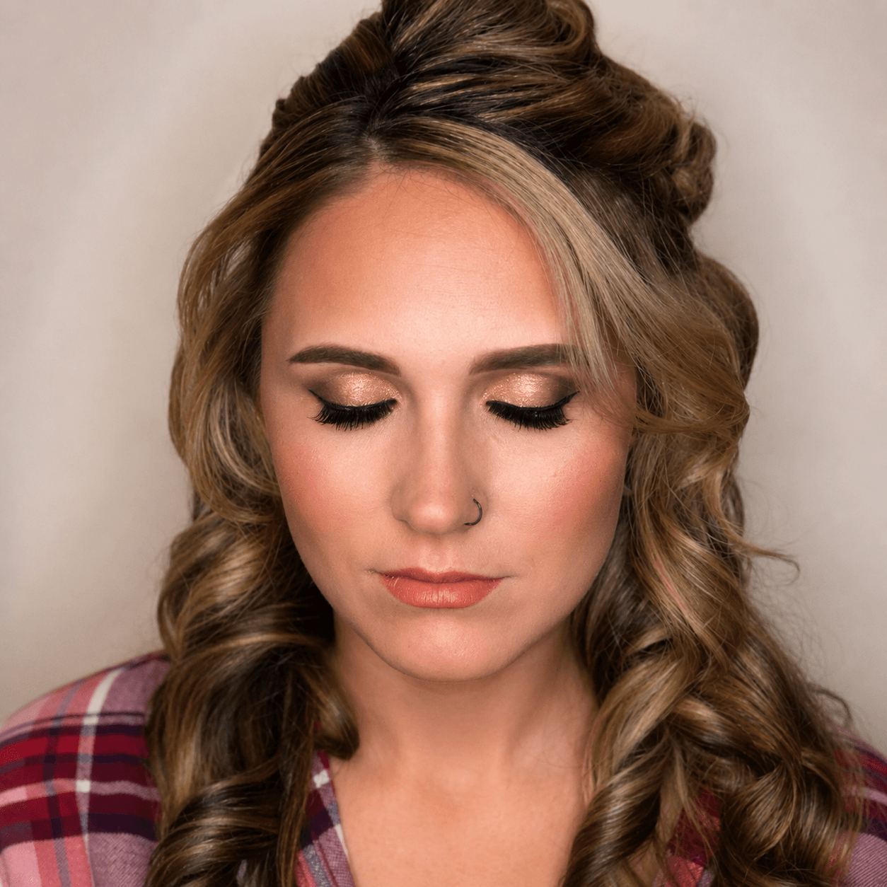 Close-up of a woman with styled wavy hair, makeup, and a nose piercing, wearing a plaid shirt, looking downward.