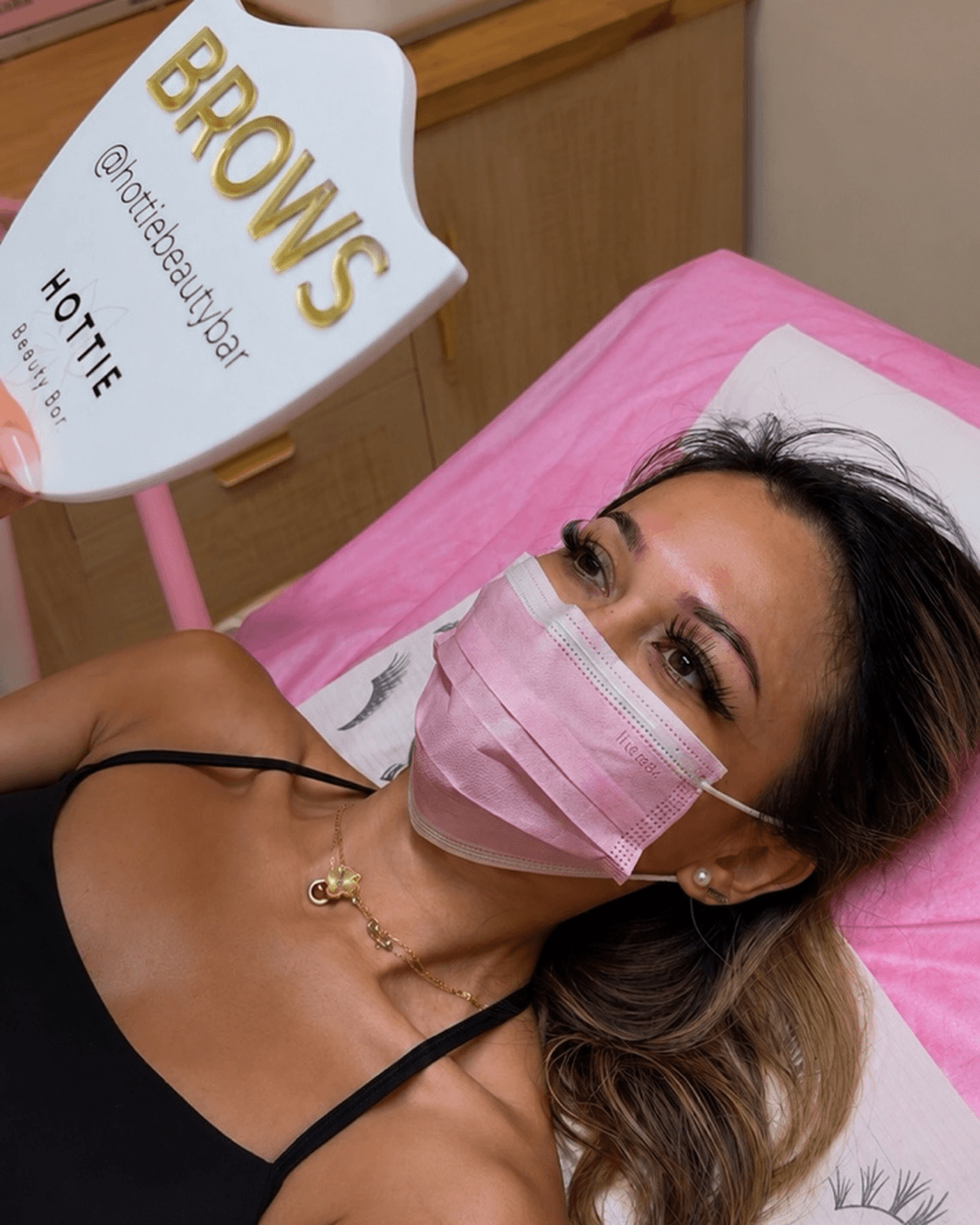 Woman lying on a pink pillow, wearing a pink face mask and black top, with a sign reading 'BROWS' nearby.