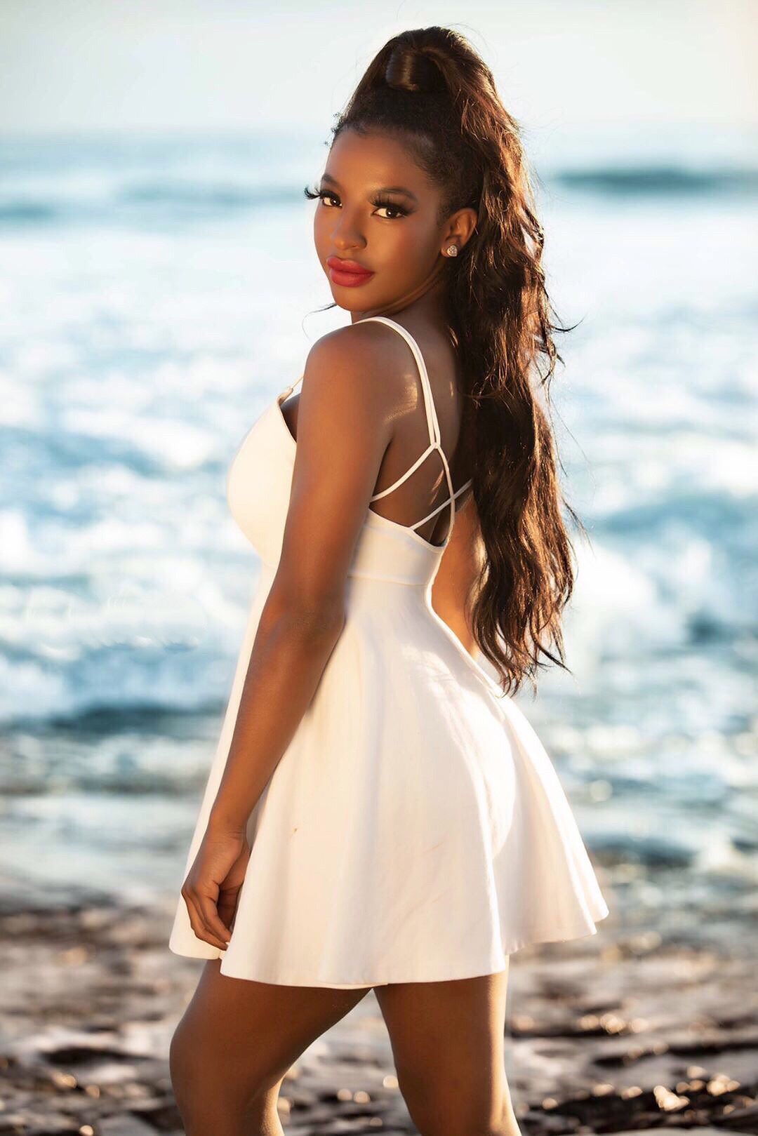 Young woman with long hair in a white dress standing on a beach with ocean waves behind her.
