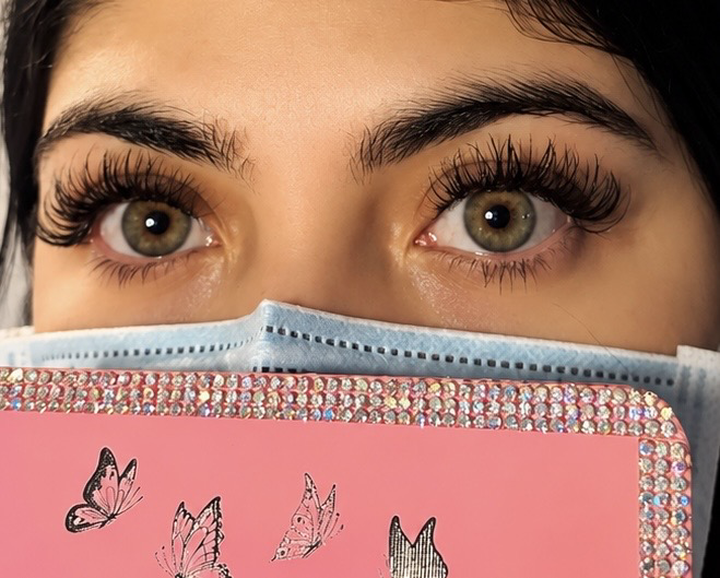 Close-up of person's eyes with long eyelashes, holding a pink notebook with butterfly illustrations and rhinestones, partially covering face.