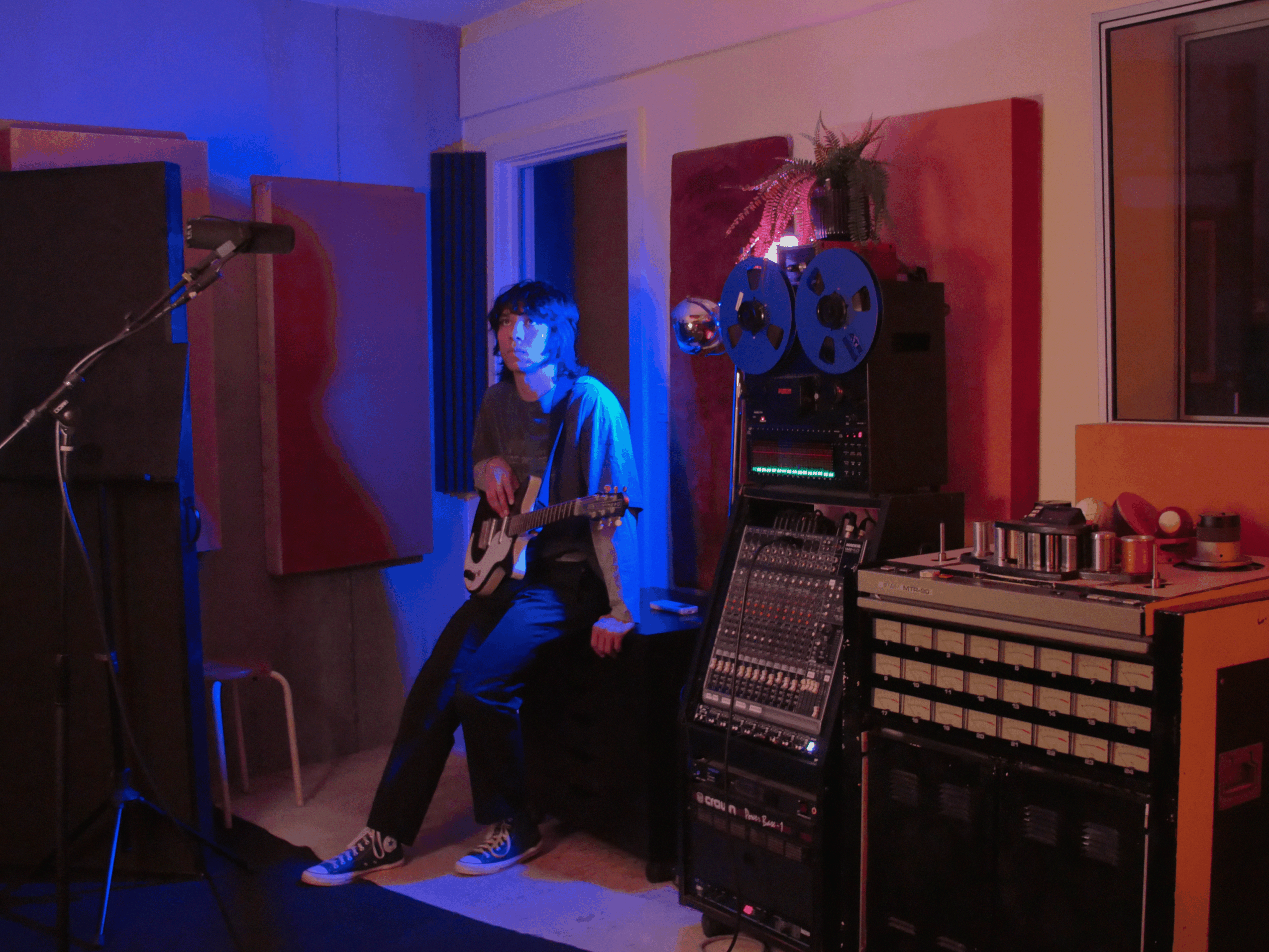 Person sitting on a box with an electric guitar in a music studio, surrounded by sound equipment and colorful wall art.