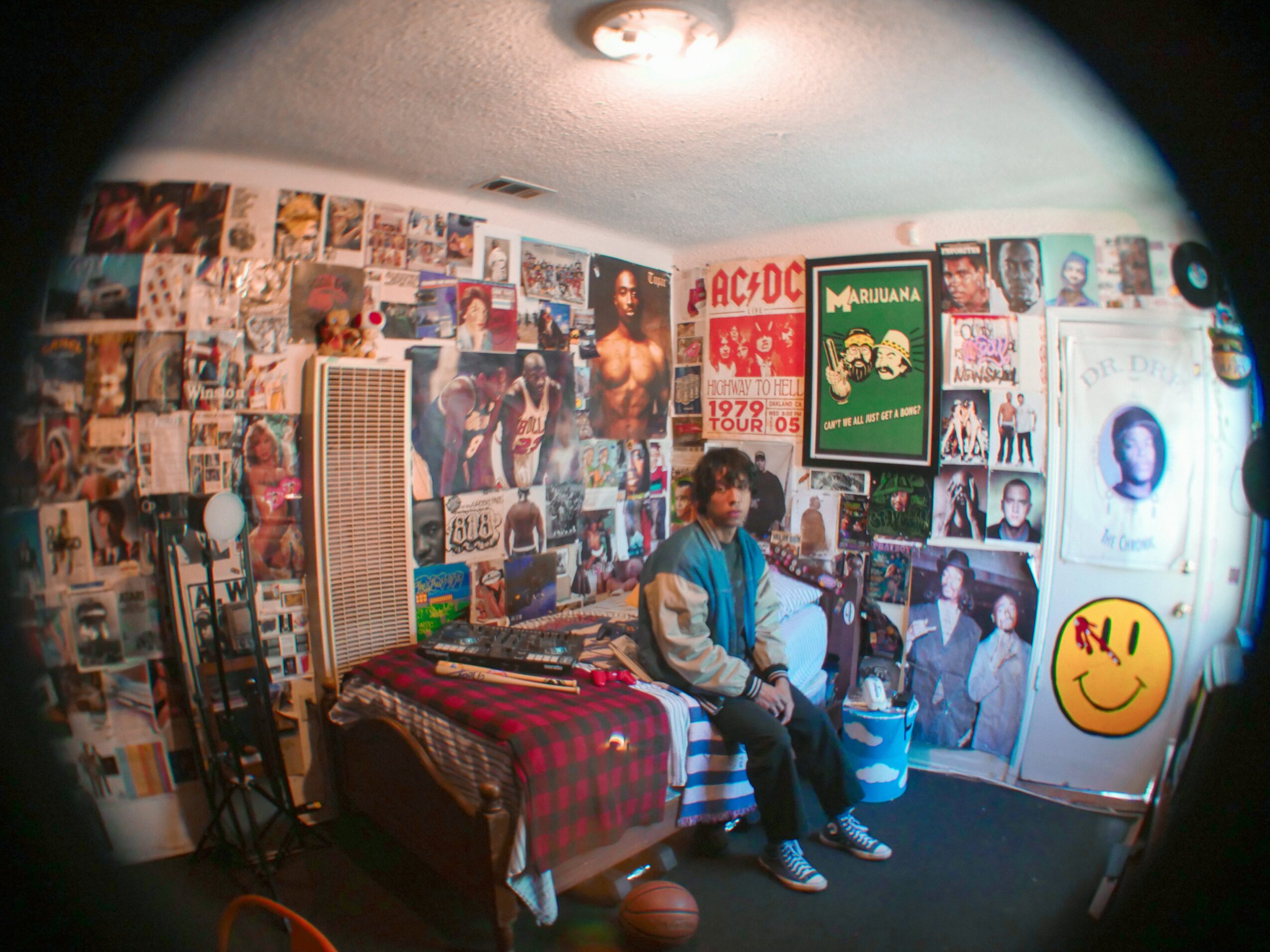 Person sitting on a bed in a room with walls covered in posters and photos, with a smiley face sticker on the wall.